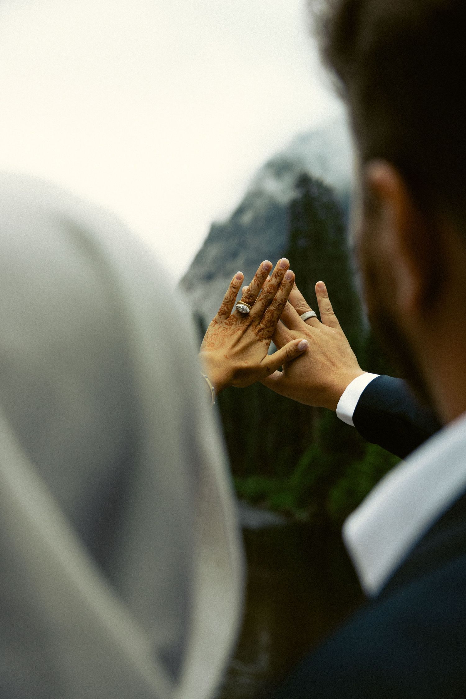 Close-up of bride and groom holding hands showing wedding rings with a misty mountain lake backdrop.