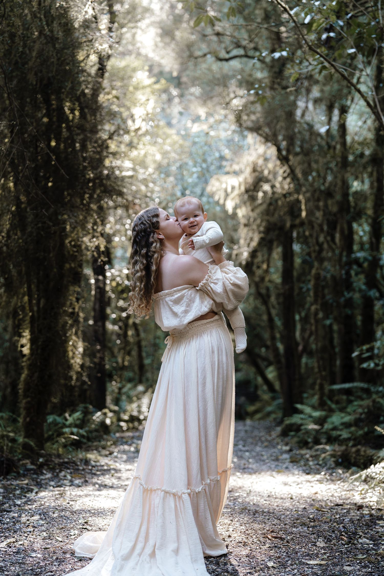 Nelson Motherhood photography photographing mother in elegant cream gown kissing baby on forest path in dappled light.