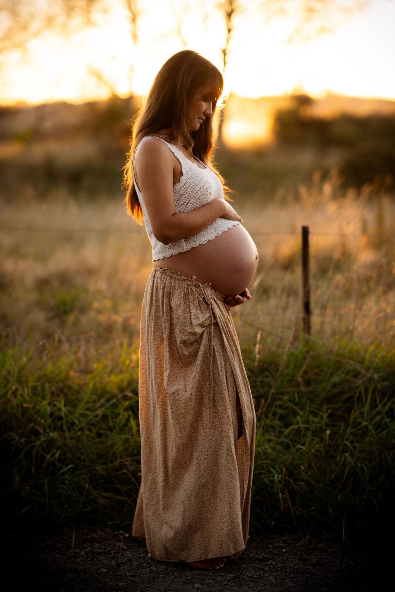Pregnant woman in white crop top and gold skirt cradling her bump during golden hour in a field. Captured by Nelson Maternity Photographer