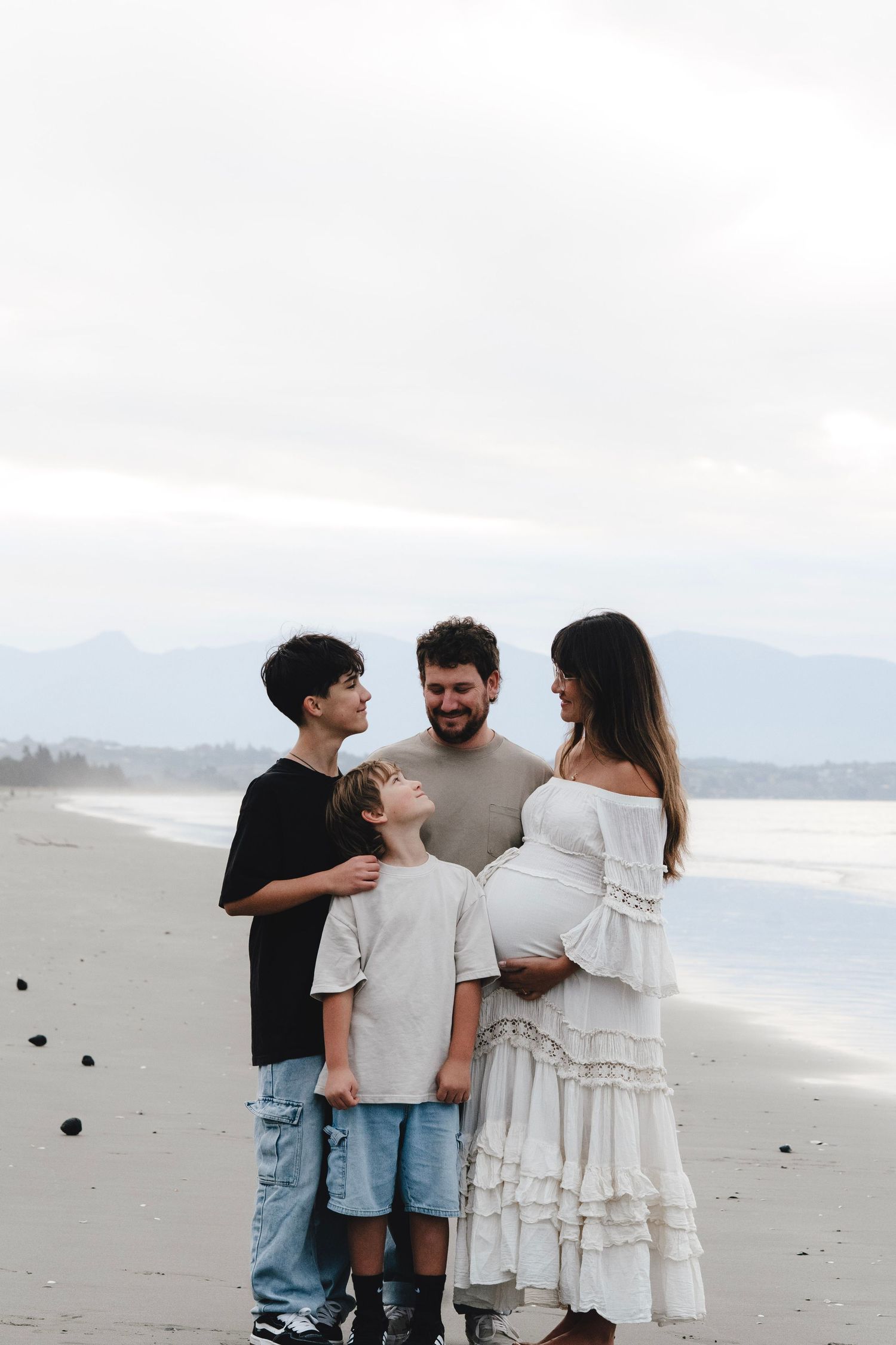 Pregnant woman in white dress with family on beach, husband and two sons smiling together. Captured by Nelson Family Photographer