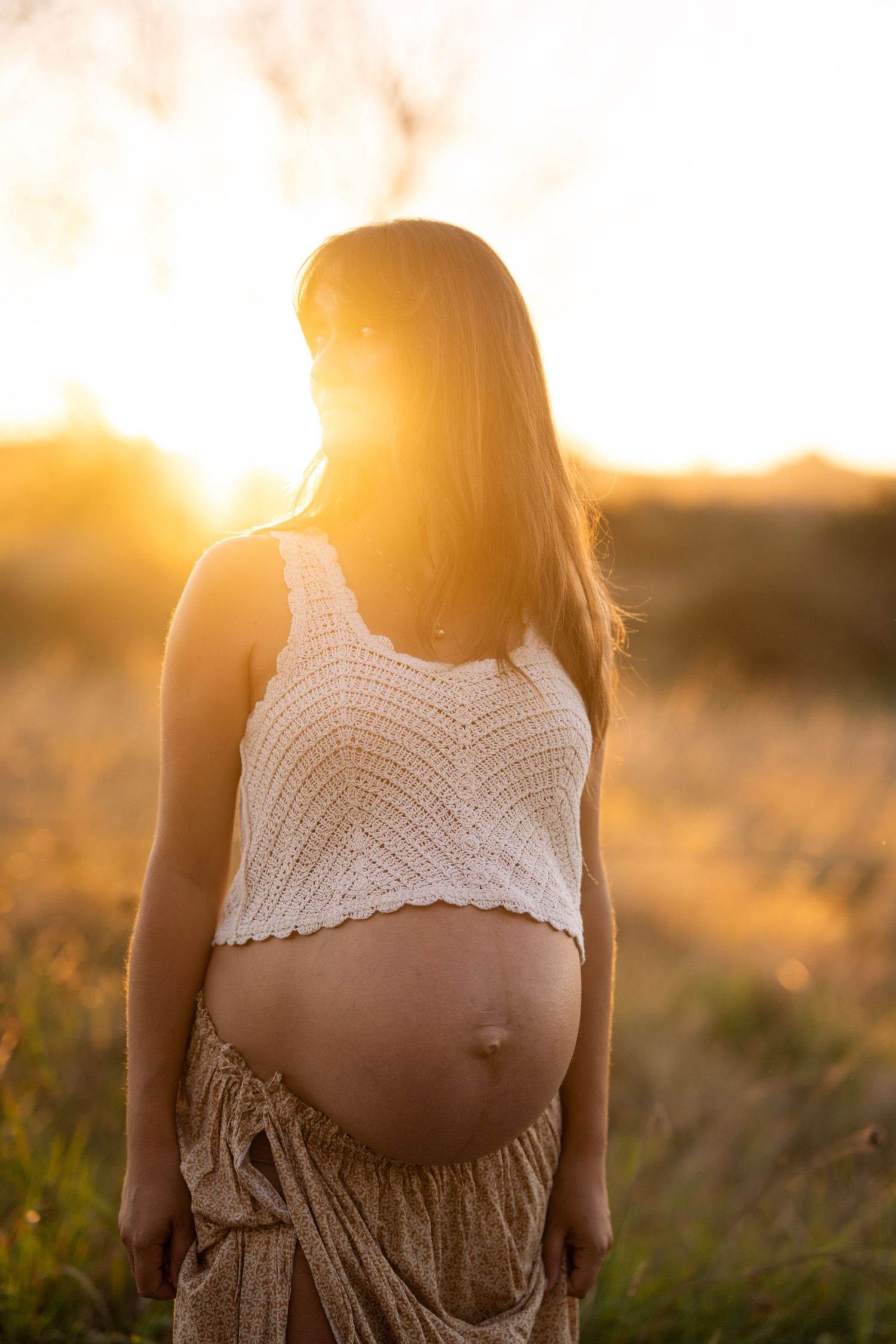 Nelson maternity photography of Pregnant woman in crochet top showing baby bump in golden sunset field glow.