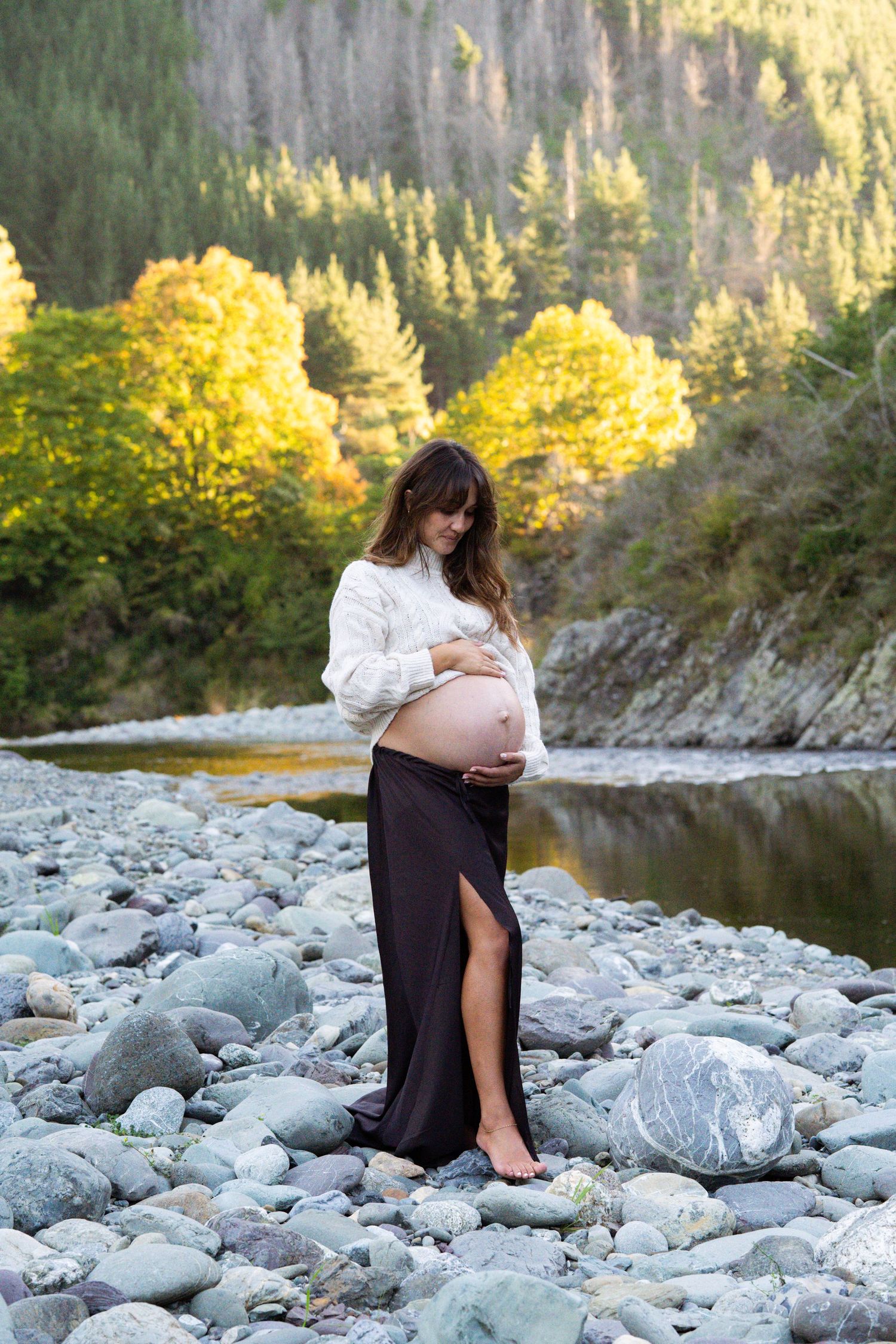 Pregnant woman in white sweater and black skirt cradles belly on rocky riverside by Nelson Maternity Photographer
