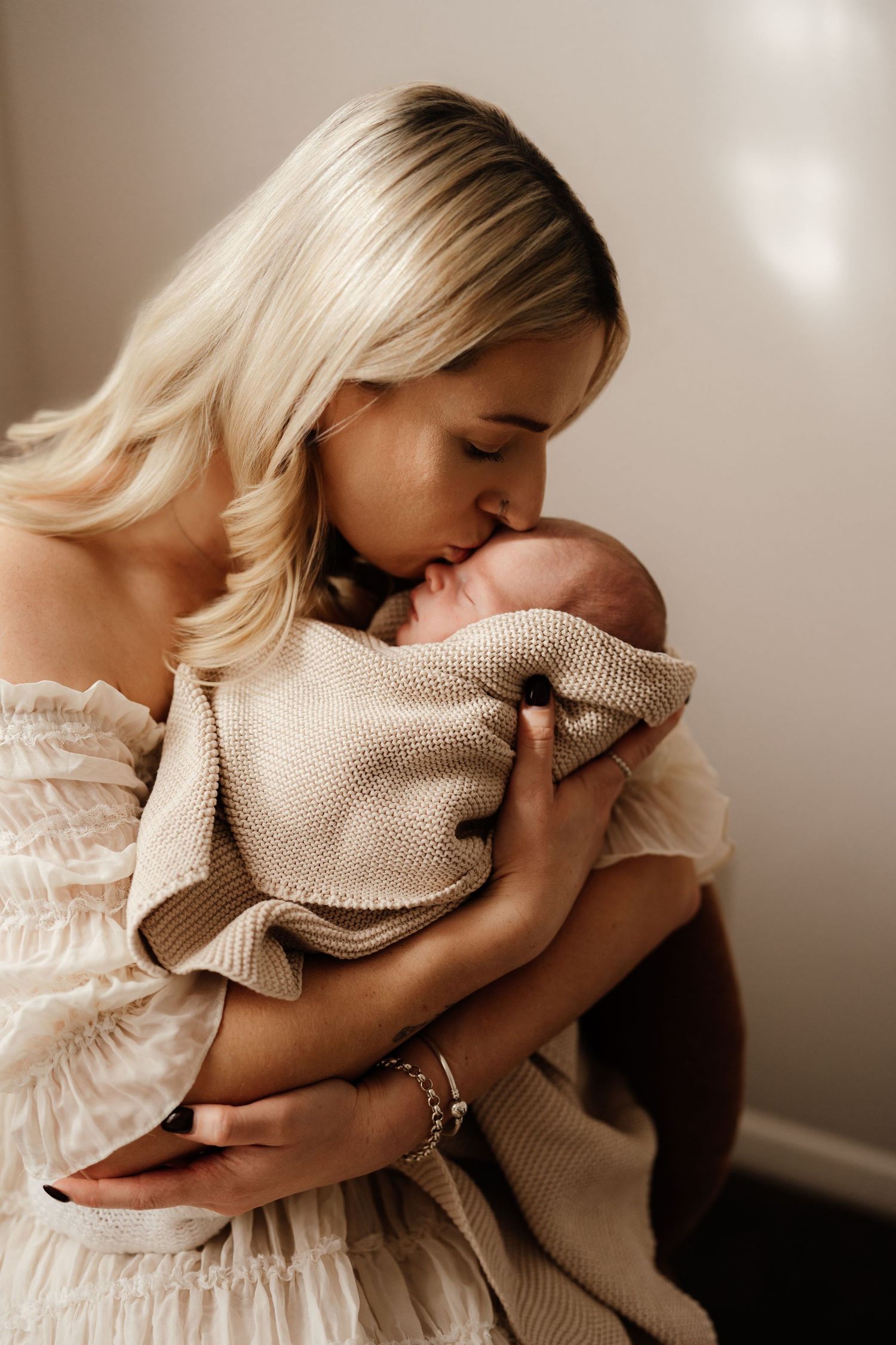 Mother in white dress tenderly kissing her newborn baby wrapped in beige knit blanket by Nelson Newborn Photographer