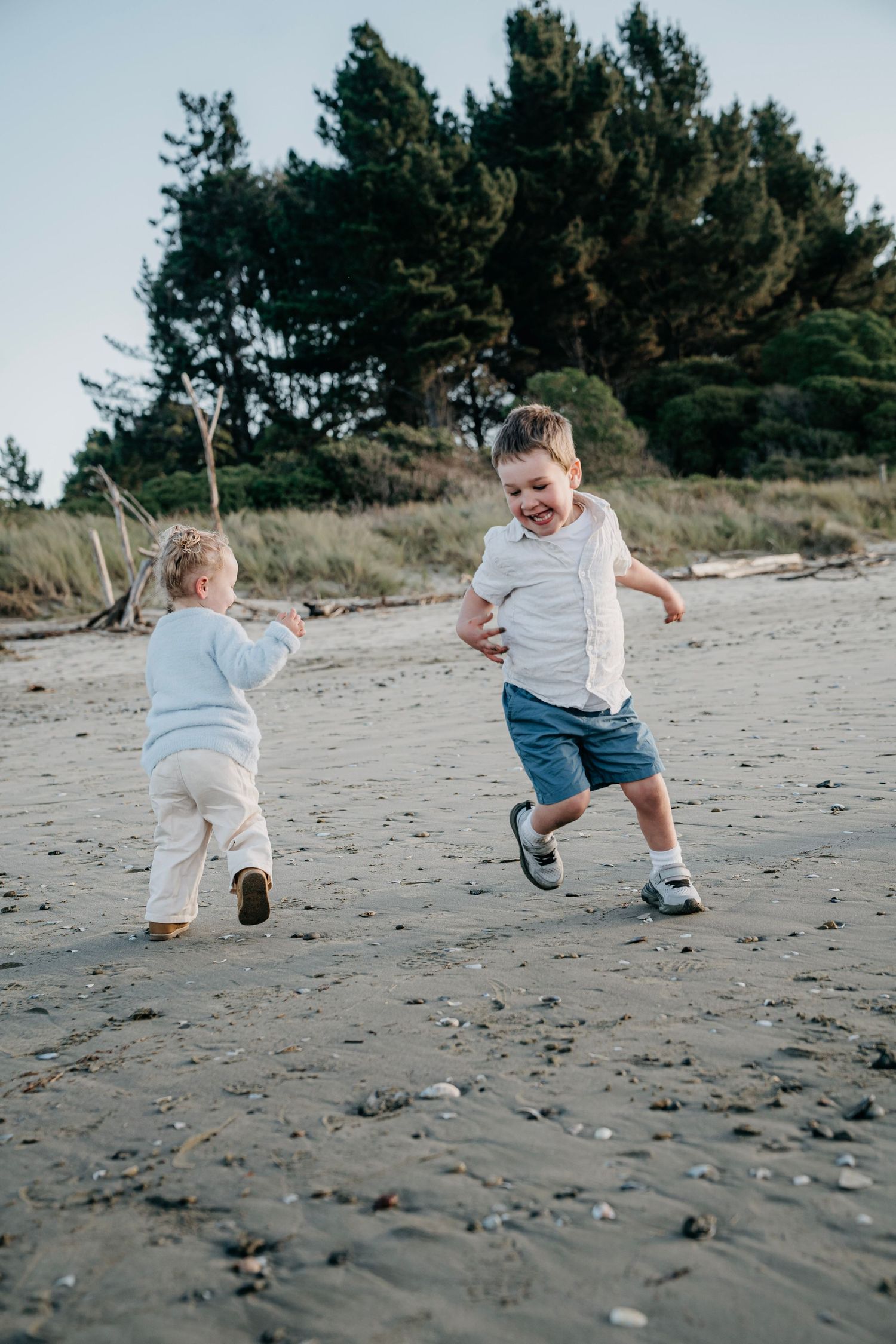 Nelson family photographer capturing Two kids laughing and playing together on a sandy beach near pine trees.