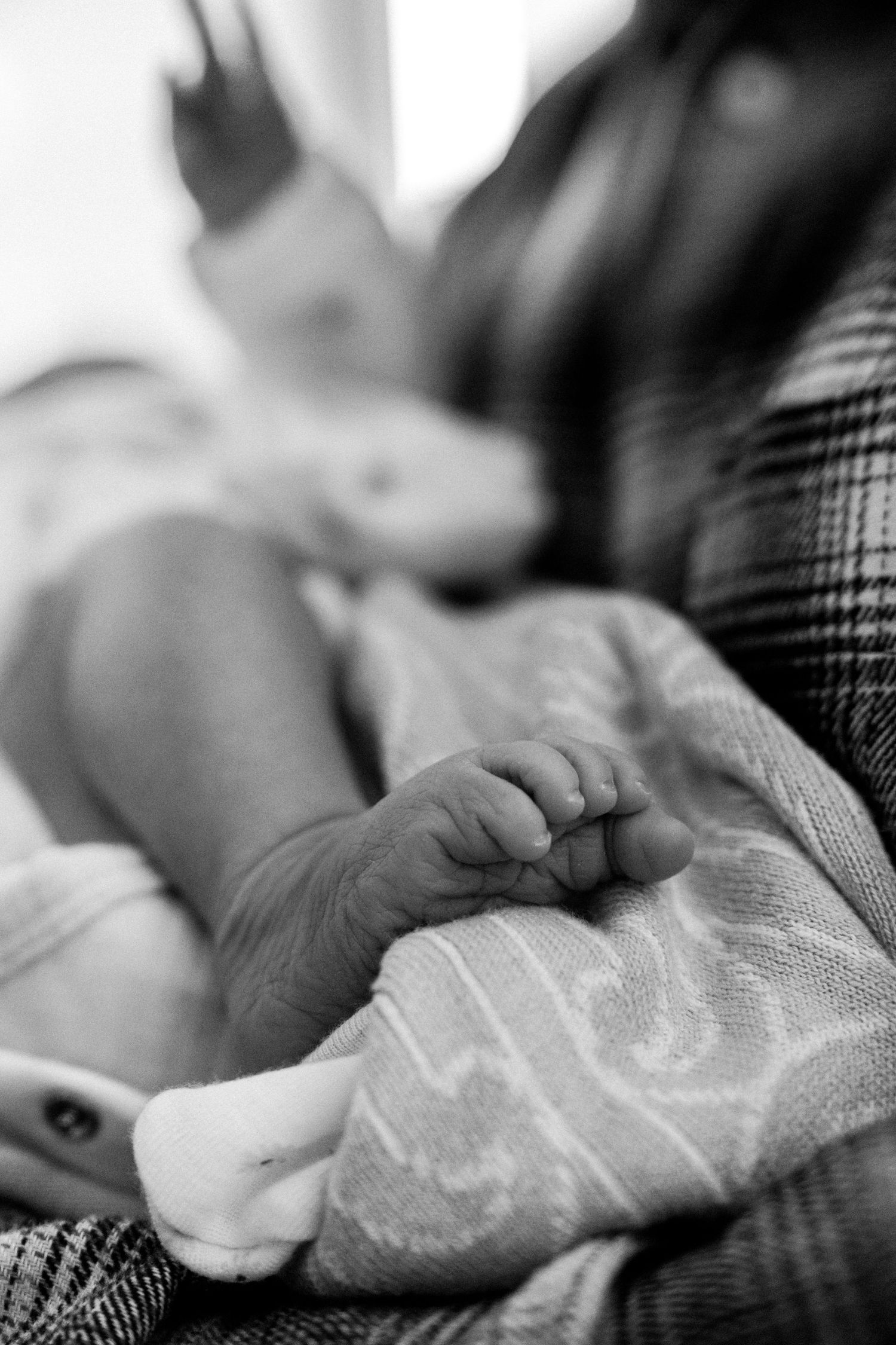 Close-up black and white photo of a newborn baby's tiny feet resting on a parent's lap in a hospital by Nelson Newborn Photographer