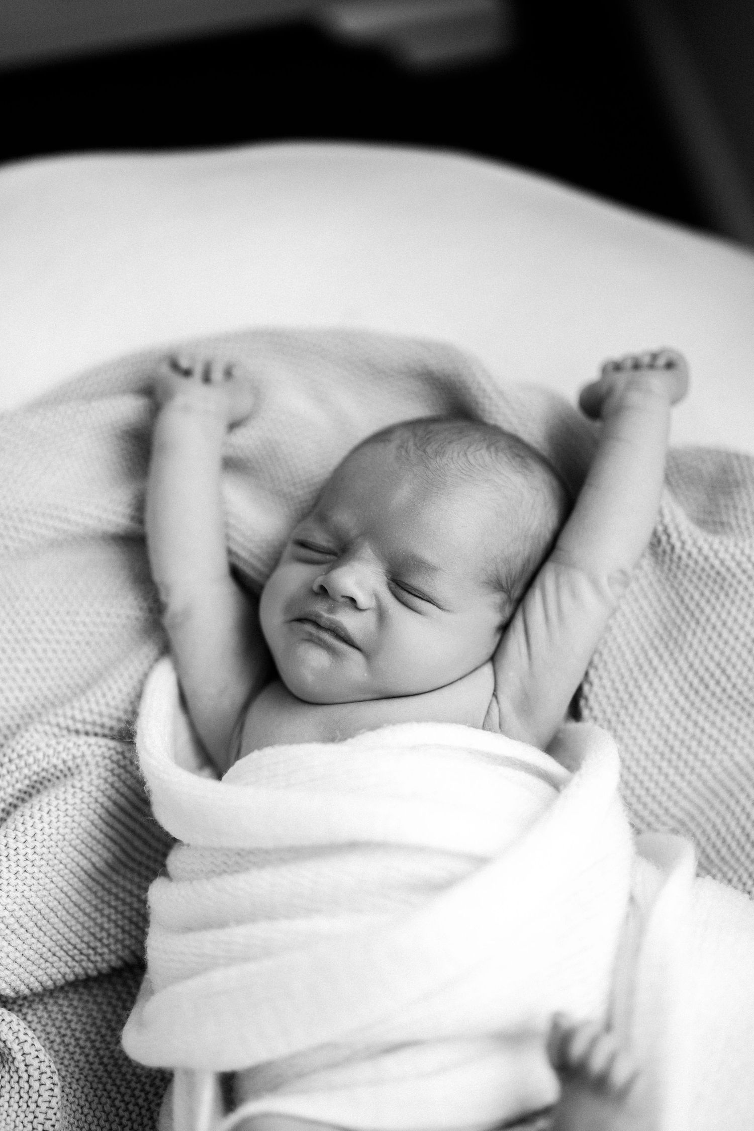 Black and white photo of newborn baby stretching with arms raised while wrapped in swaddle  by Nelson Newborn Photographer