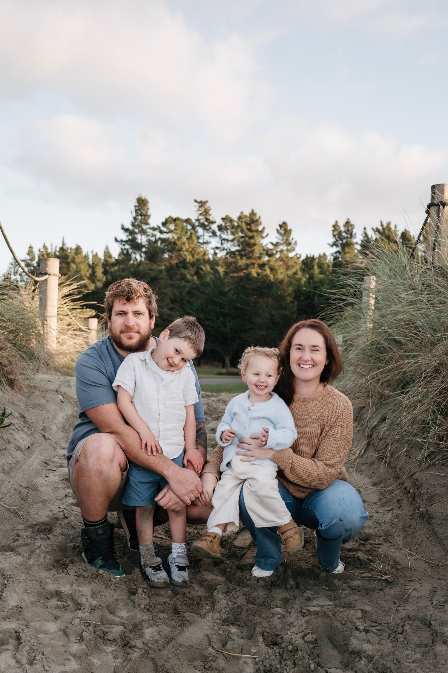Nelson family photography of Happy family of four posing together in sandy beach dunes with pine trees.