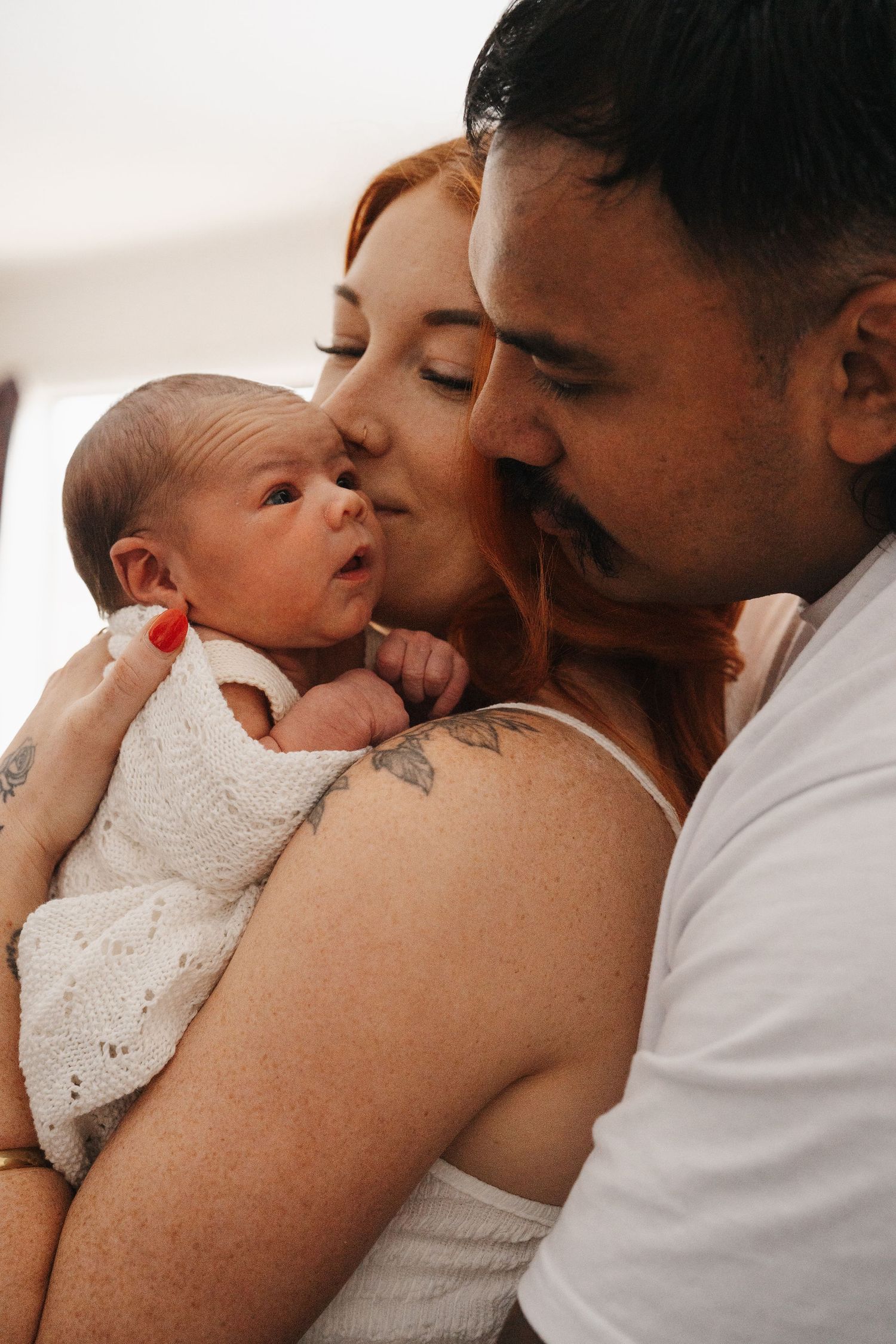 Nelson newborn photography of New parents tenderly kissing their newborn baby wrapped in white knit blanket.