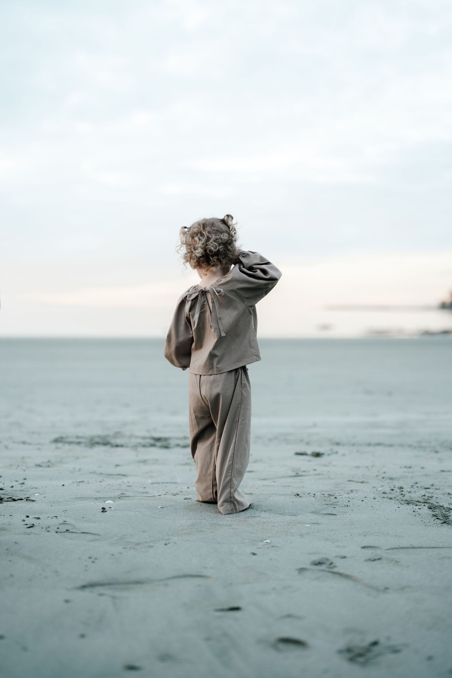 Nelson family photography of Curly-haired toddler in grey outfit standing alone on vast sandy beach at dusk.