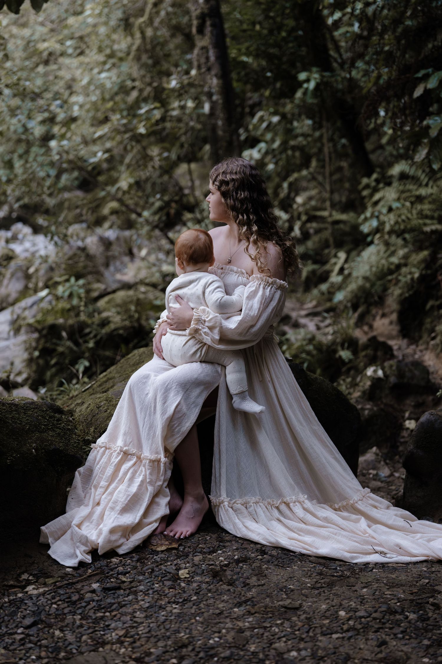 Woman in elegant white off-shoulder gown sitting on mossy rocks in a lush forest holding her baby. Captured by Nelson Photographer