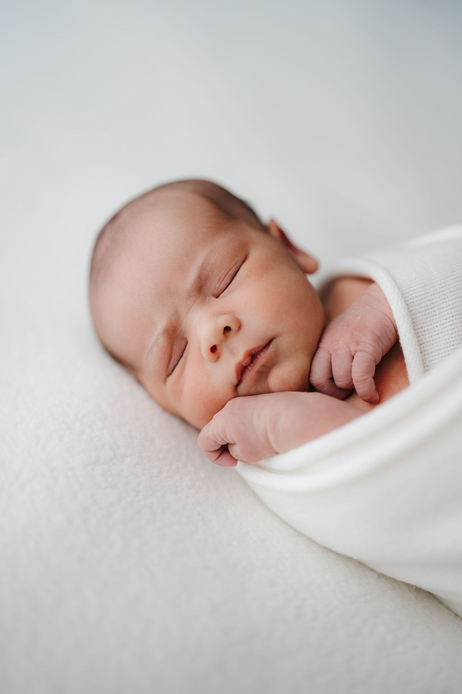 Peaceful newborn baby sleeping soundly wrapped in a white swaddle on a soft blanket  by Nelson Newborn Photographer