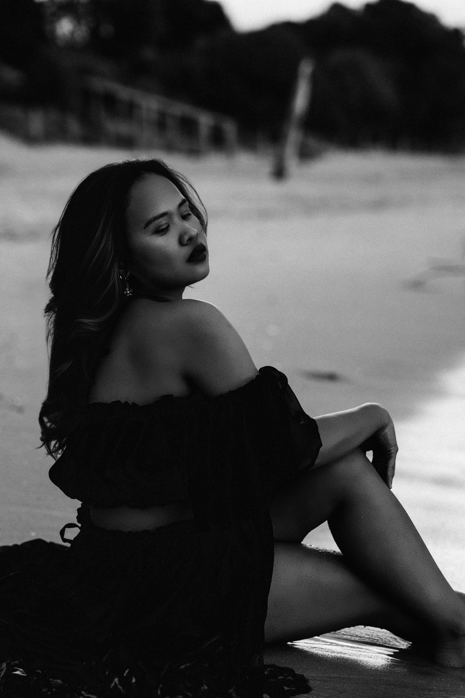 Black and white portrait of a woman in off-shoulder black dress sitting on a beach at dusk. Captured by Nelson Photographer