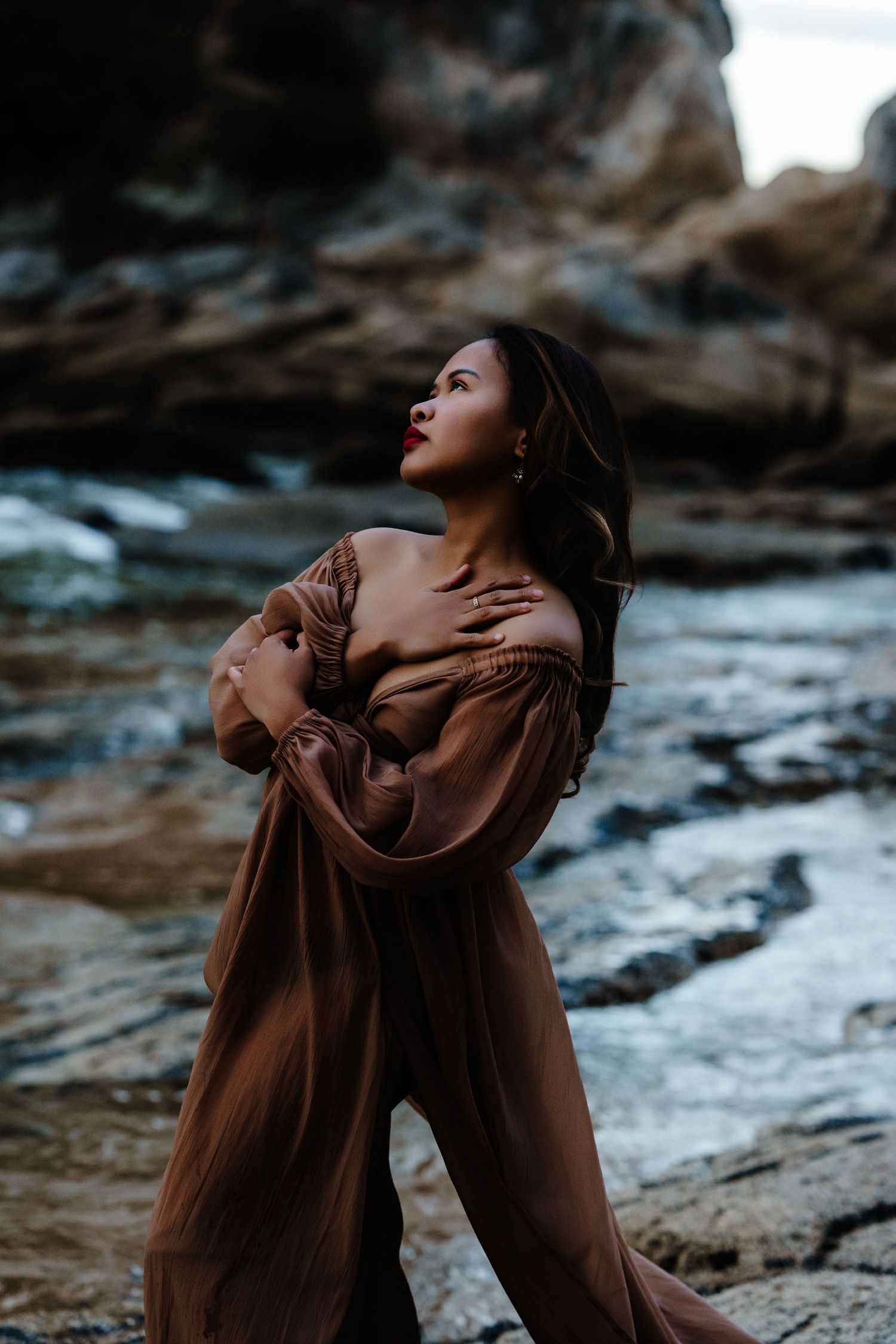 Nelson Motherhood Photography of Elegant woman in flowing brown off-shoulder dress posing dramatically among coastal rocks.