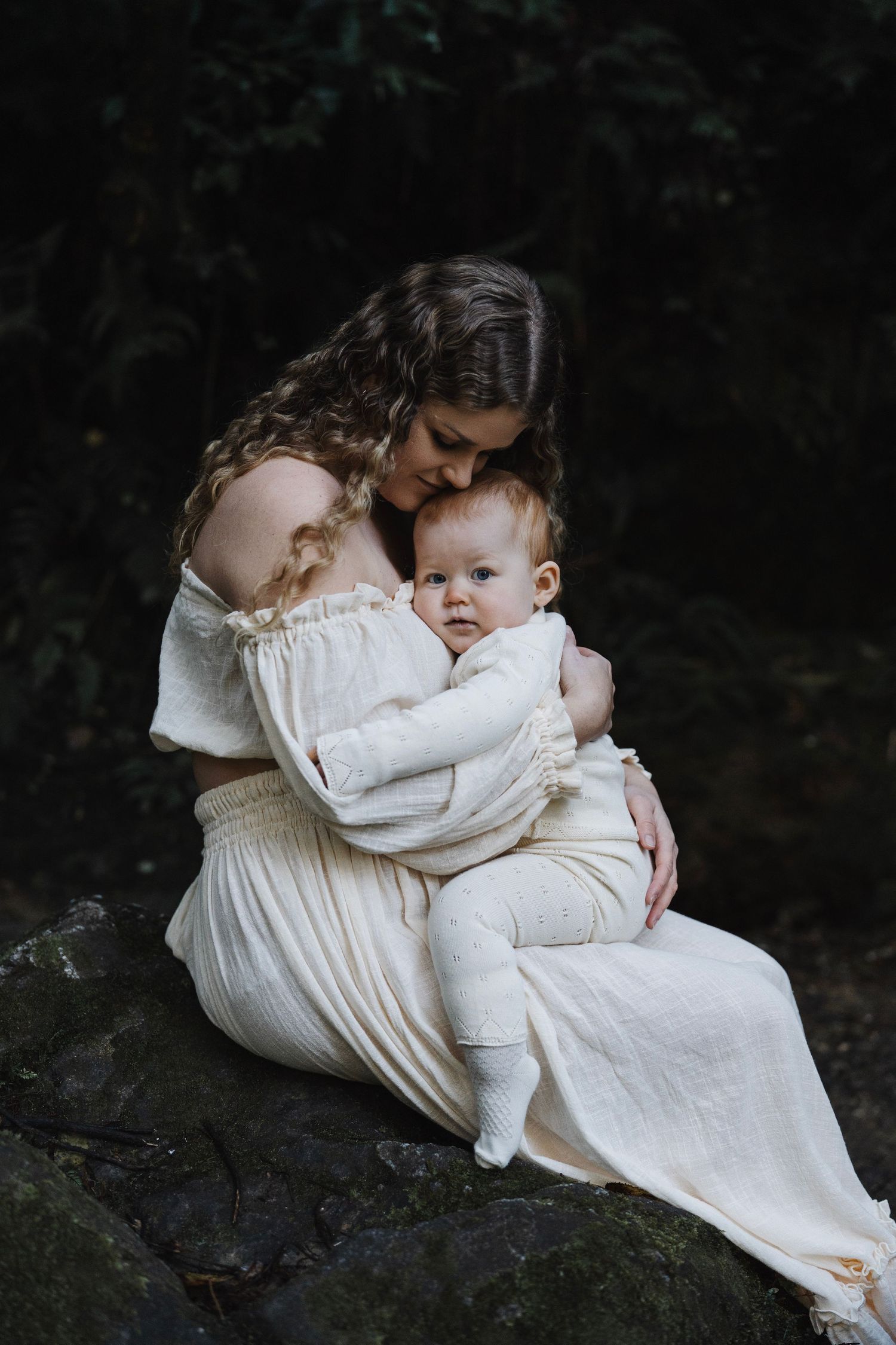 Mother in white dress sitting on rocks in dark forest, tenderly embracing her wide-eyed baby. Captured by Nelson Motherhood Photographer