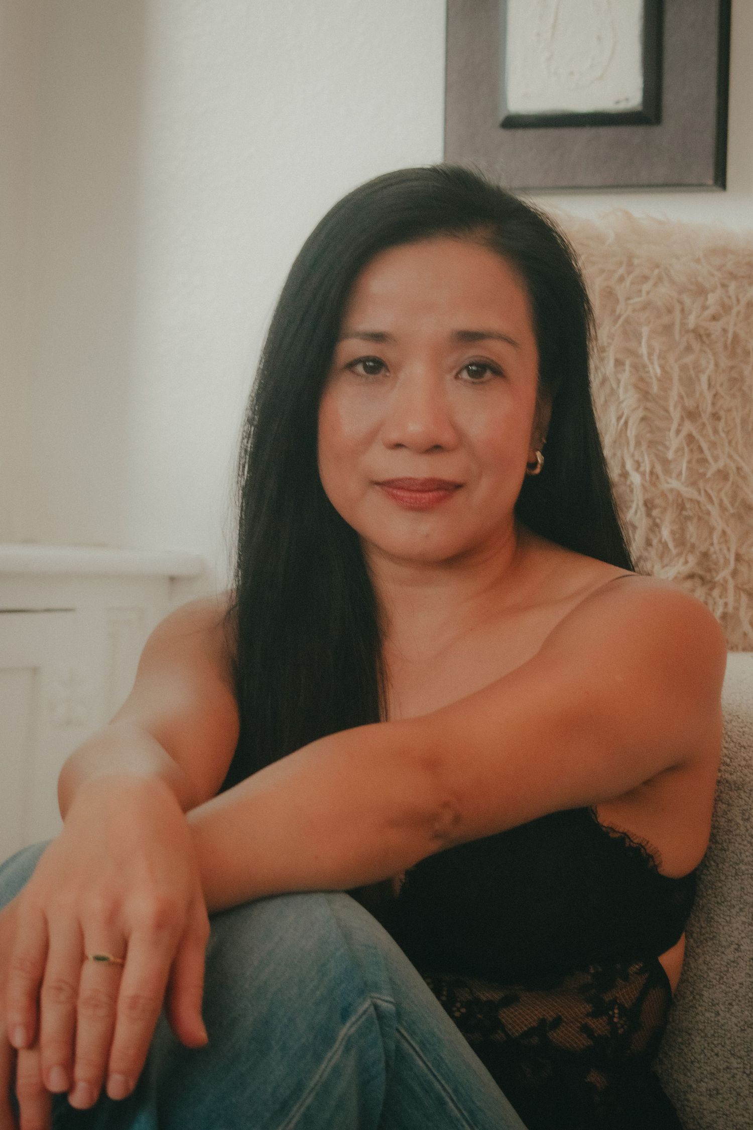 Woman with long dark hair sitting relaxed in a chair, wearing a black top and jeans, smiling warmly.
