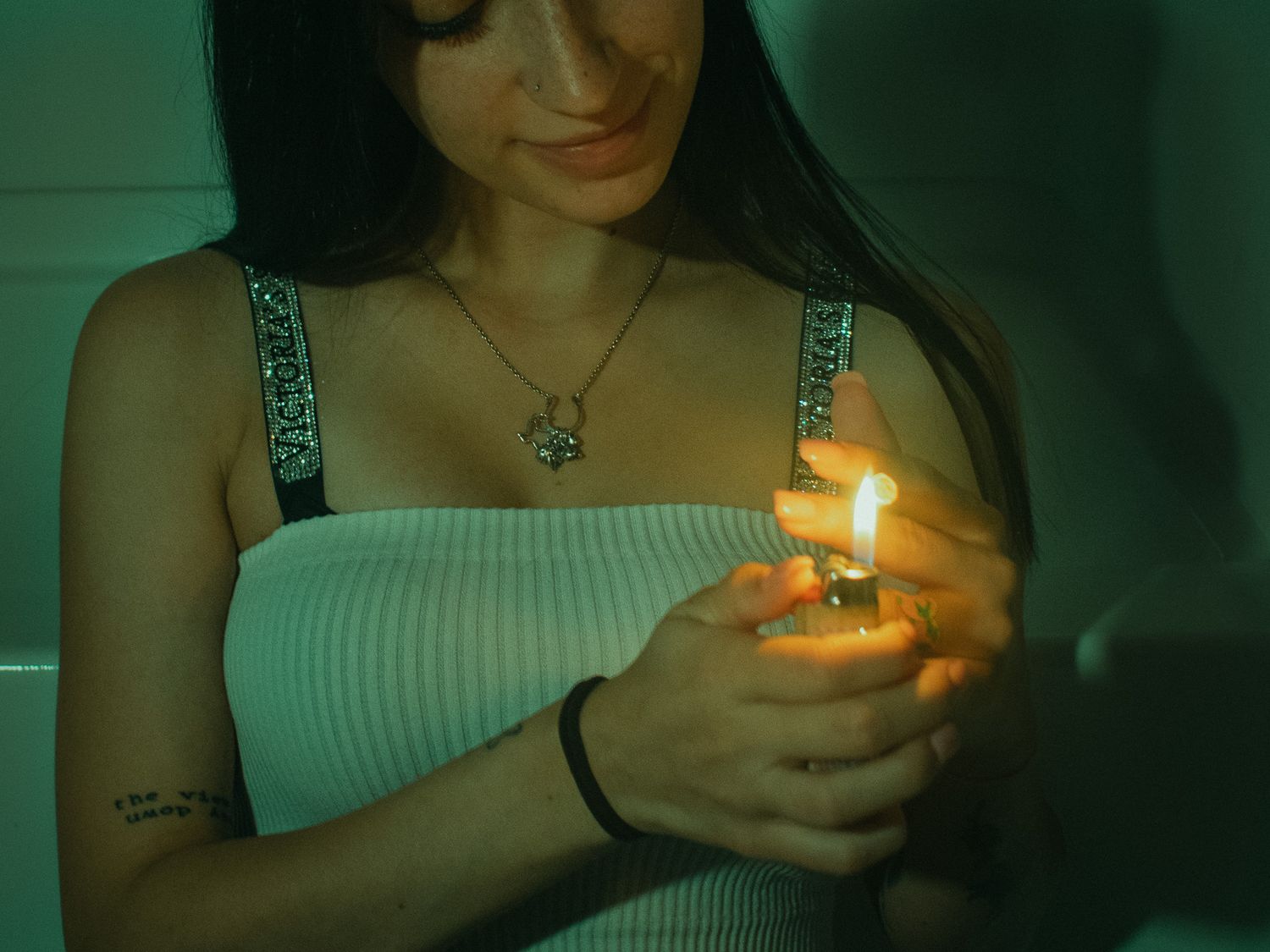 Young woman with dark hair lighting a flame with a lighter, wearing a white top and necklace indoors.