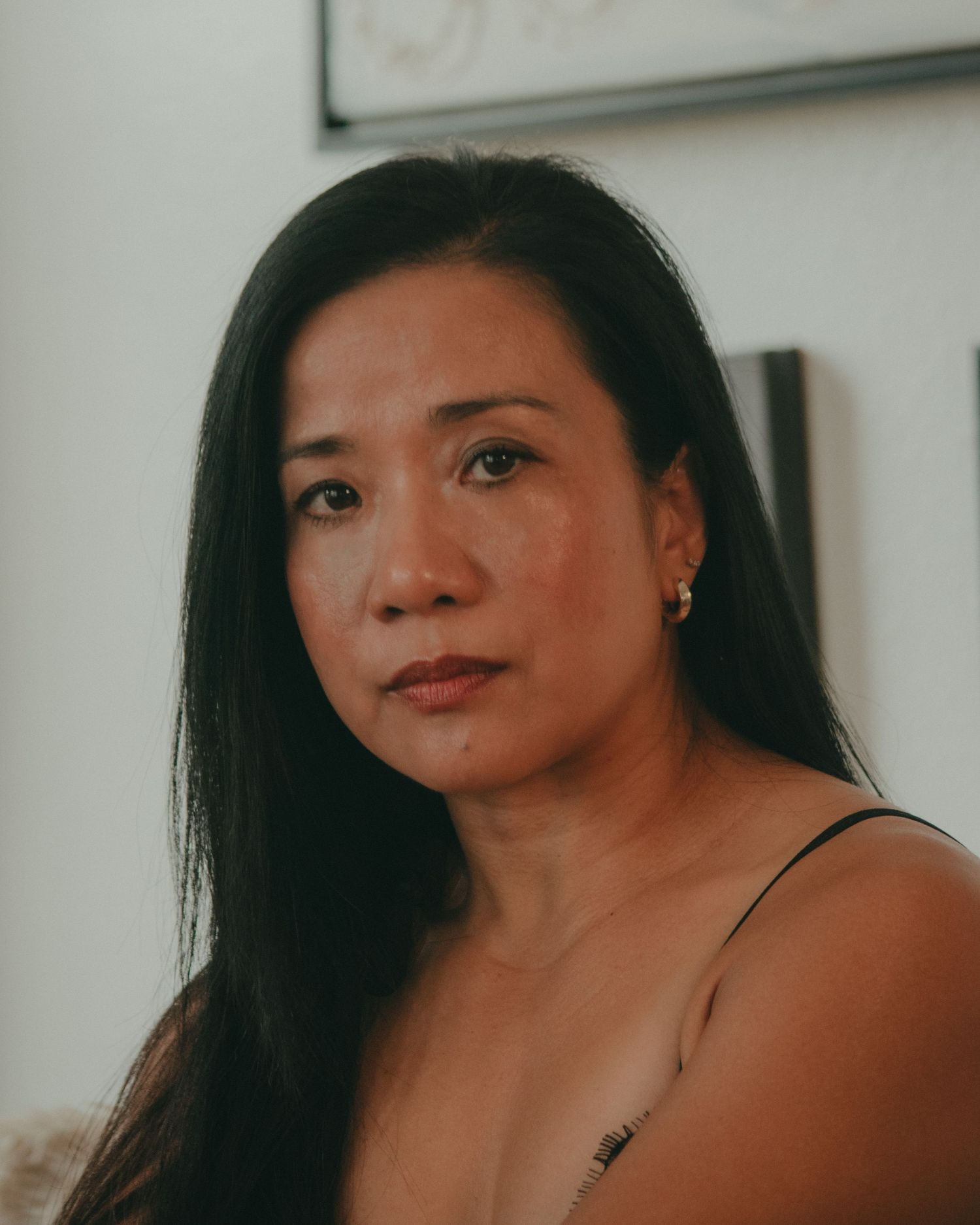 A Southeast Asian woman with long dark hair and small earrings poses indoors with a serious expression.