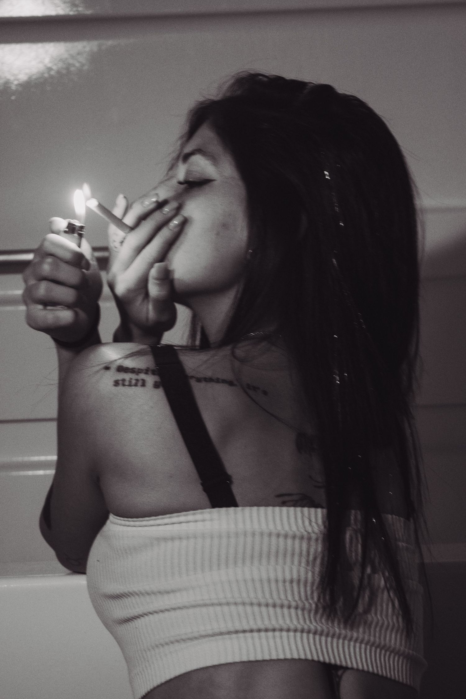 Black and white photo of young woman with tattoos lighting a cigarette indoors.