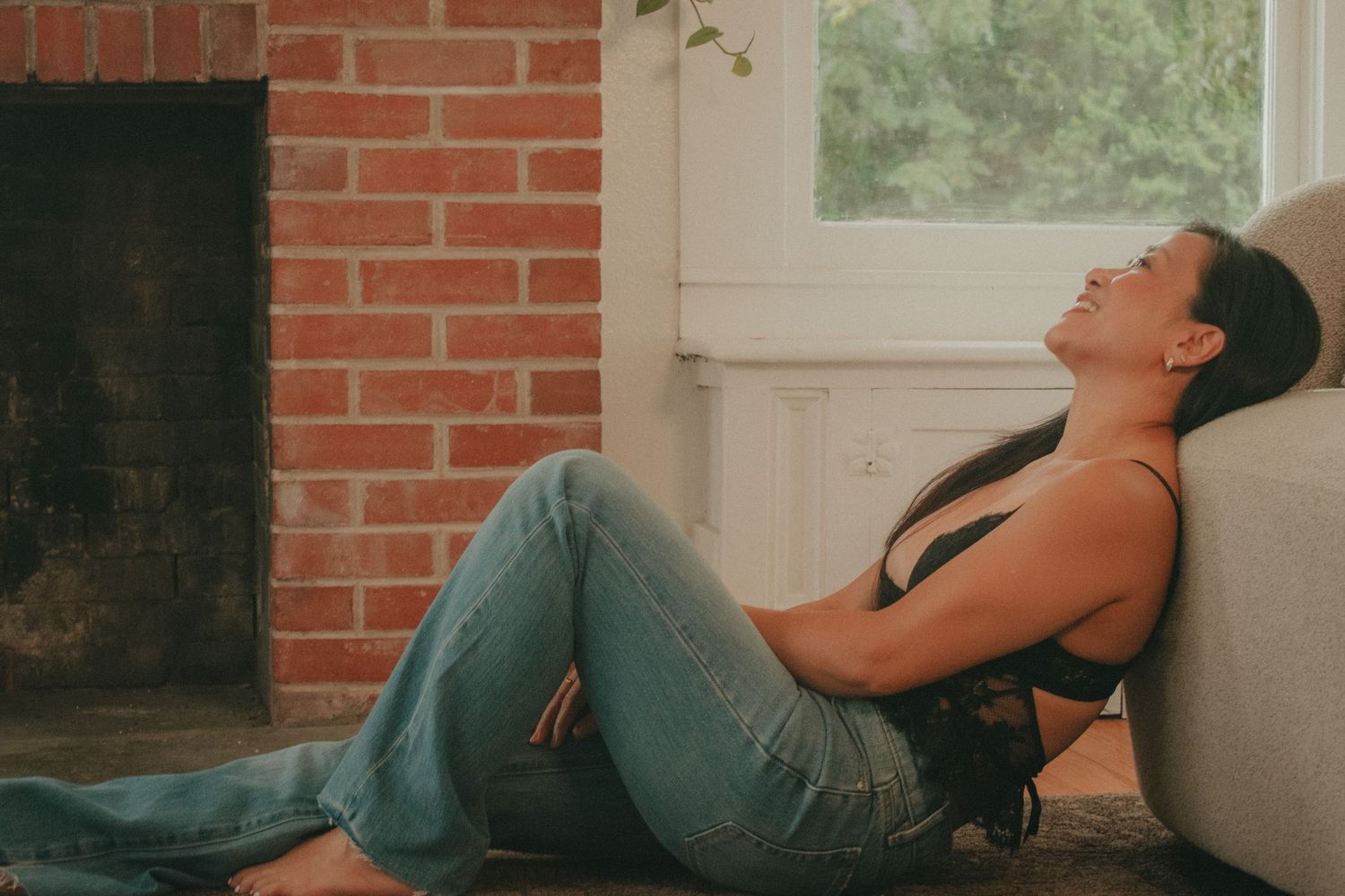 Woman with dark hair leaning back against a sofa near a brick fireplace in a relaxed pose.