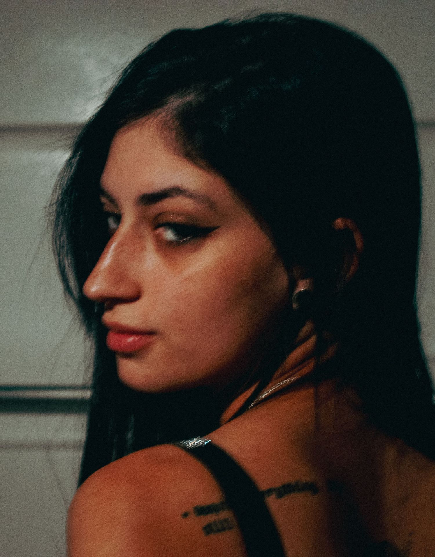 Young woman with dark hair and tattoos on her shoulder glances back over her shoulder in a moody portrait.