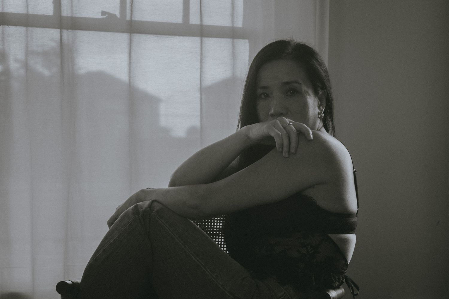 Black and white photo of a young woman sitting pensively by a curtained window with arms crossed.