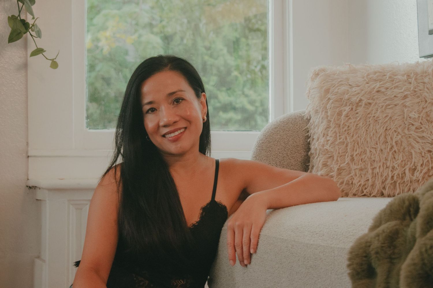 Smiling woman with long dark hair sitting relaxed on a cream sofa near a bright window indoors.