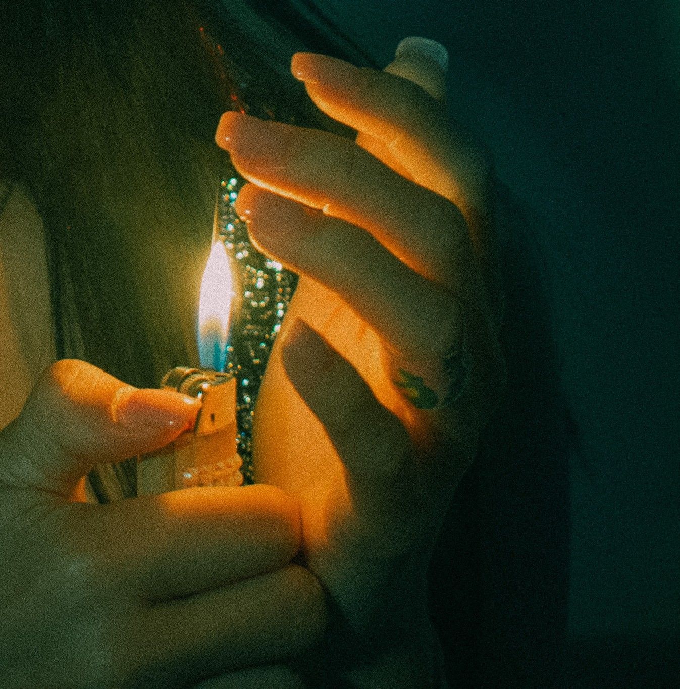 Close-up of hands holding a lit lighter, with green nail polish visible, casting a warm glow in dim lighting.