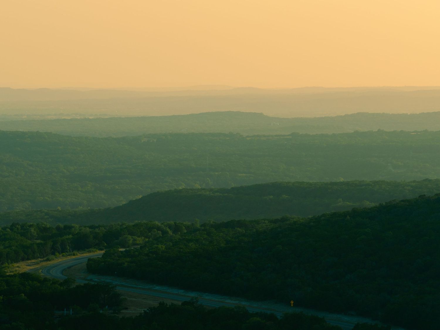 Aerial view of rolling green hills and a winding road at golden hour in a hazy landscape.