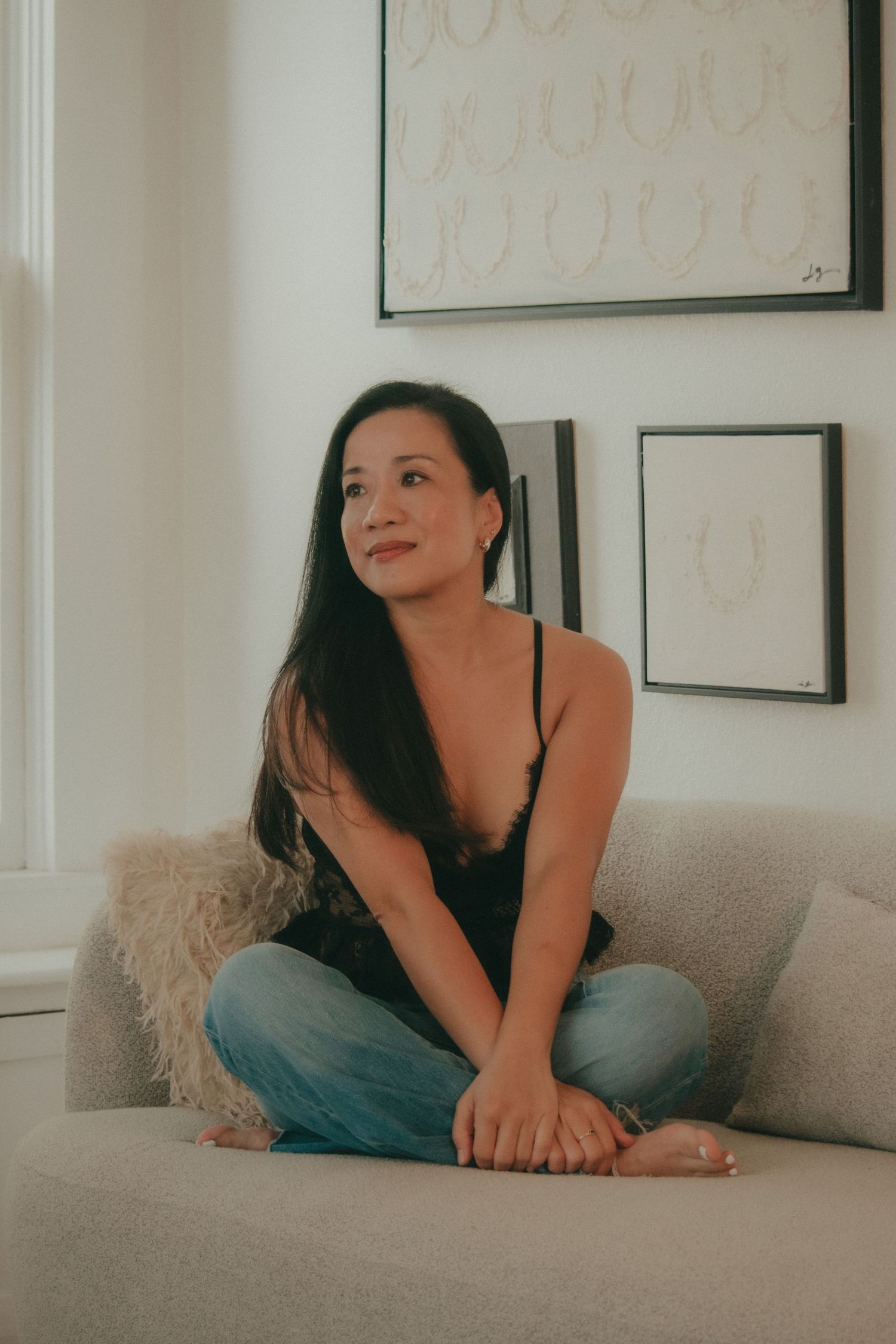 Woman in black camisole and jeans sitting cross-legged on a beige sofa, smiling softly in a bright modern living room.