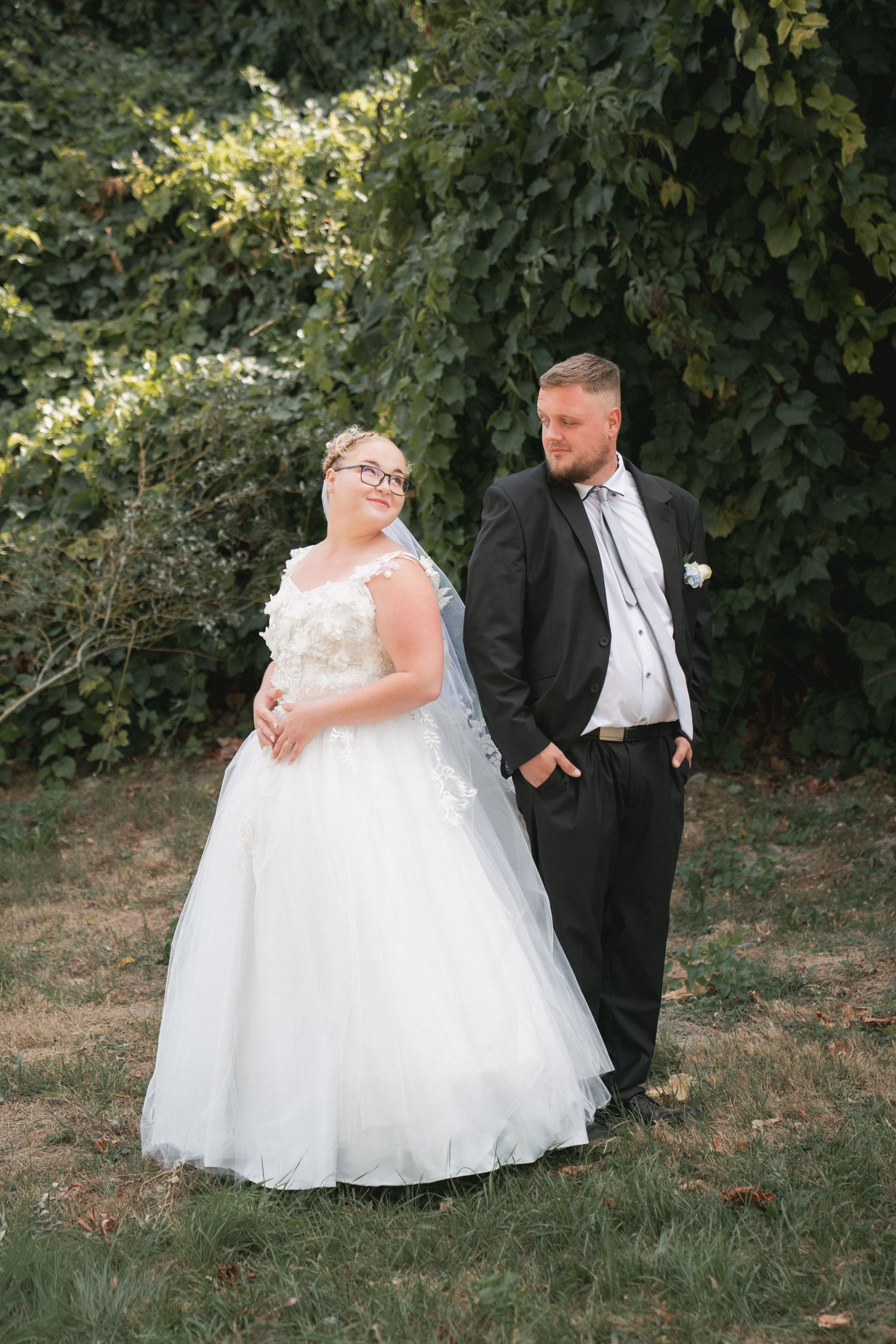 Vőlegény és menyasszony egymásnak háttal állva egy erdőben miközben egymásra pillantanak természetes fényekkel. Őszinte esküvői pillanat.