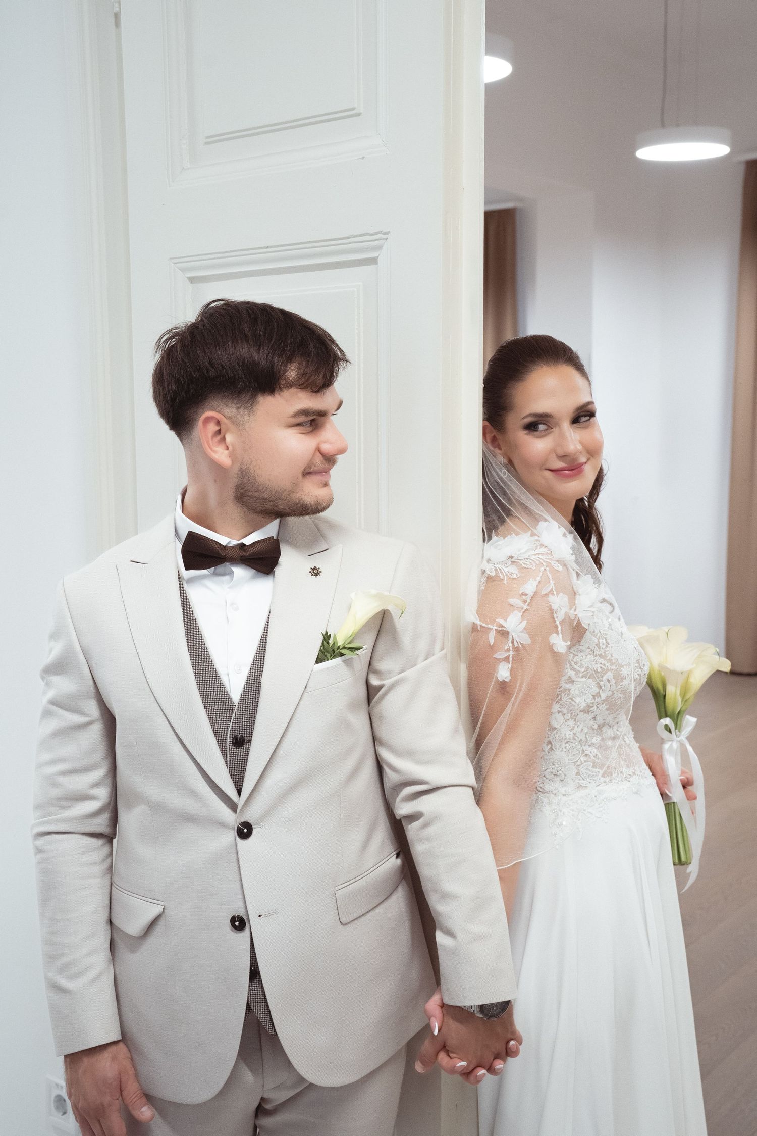 Bencze Máté fotós által készített fotó, ahol a menyasszony és a vőlegény egymás kezét fogva állnak egy szobában.