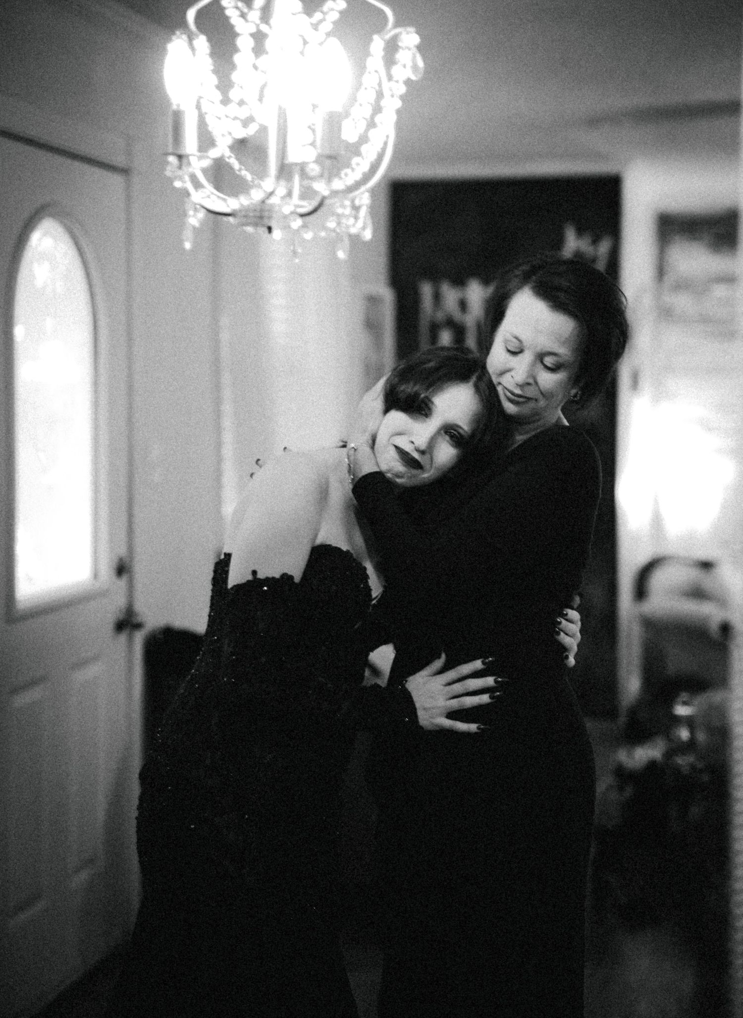 Black and white photo of bride in black gown sharing warm embrace with woman under chandelier.