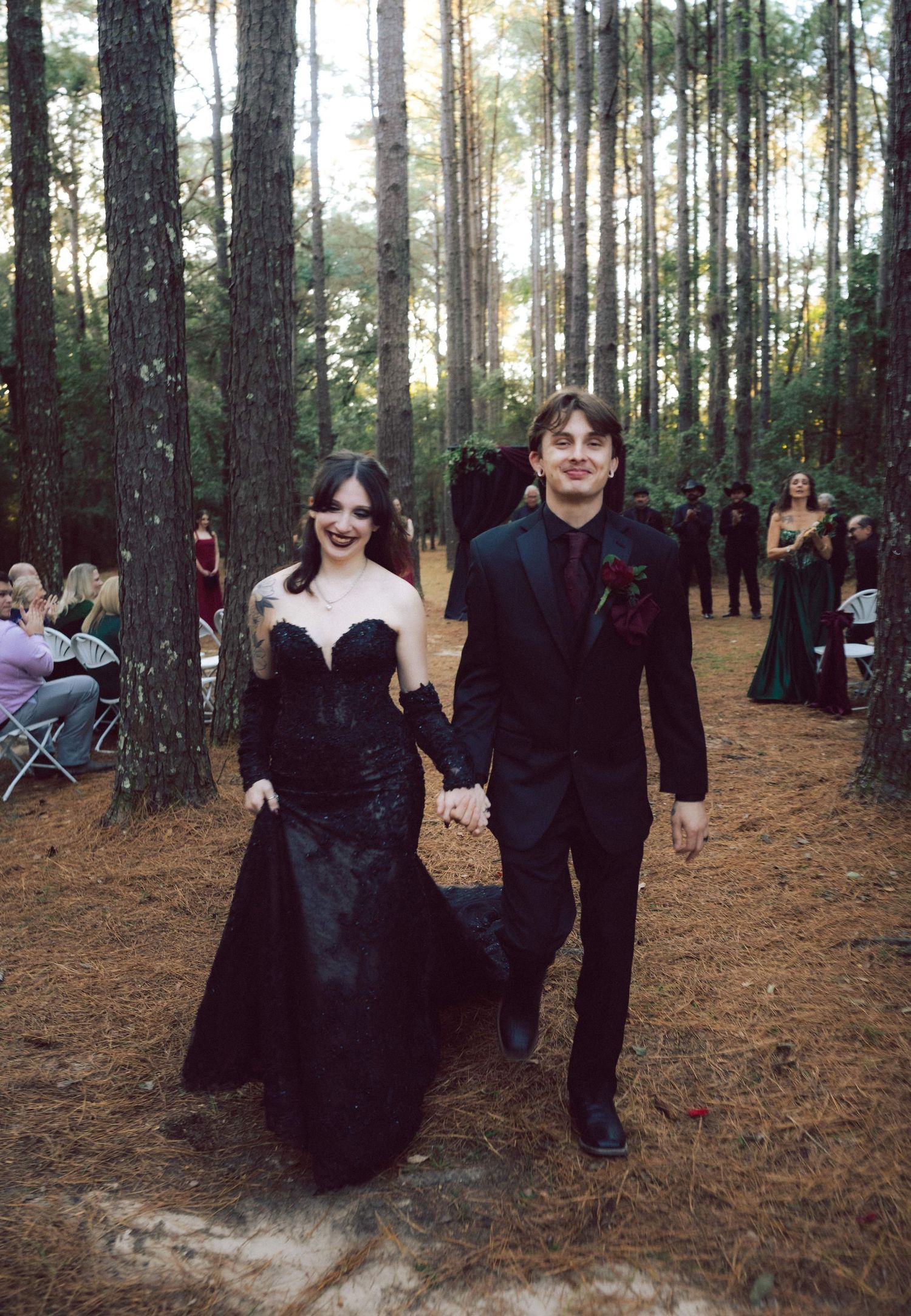 Couple in matching black wedding attire walking hand-in-hand through a pine forest ceremony aisle.