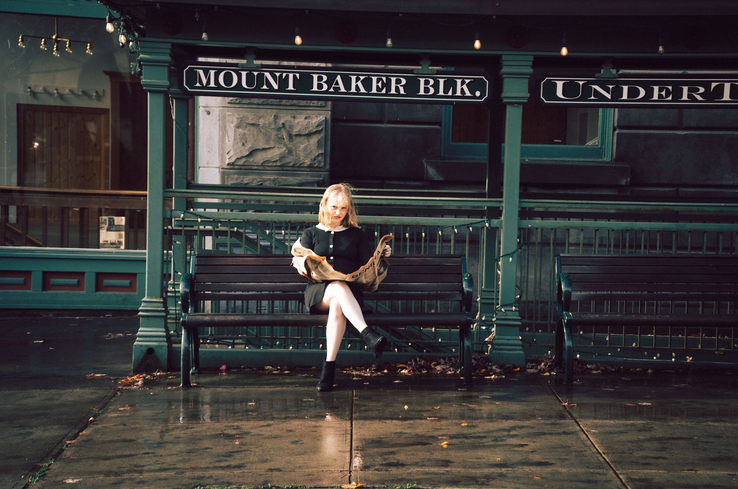 Blonde woman sitting on a bench outside Mount Baker BLK building at night, holding a dark clutch purse.