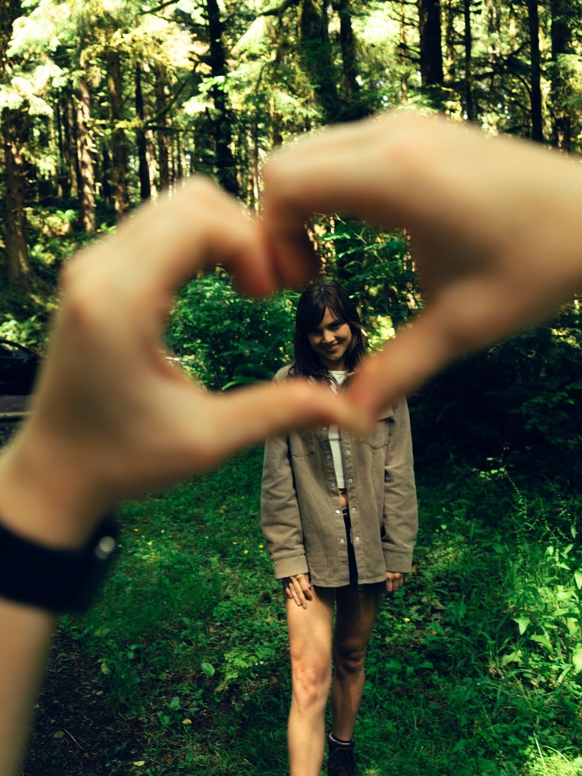 Person framing their partner with a heart hand gesture in a lush green forest setting.