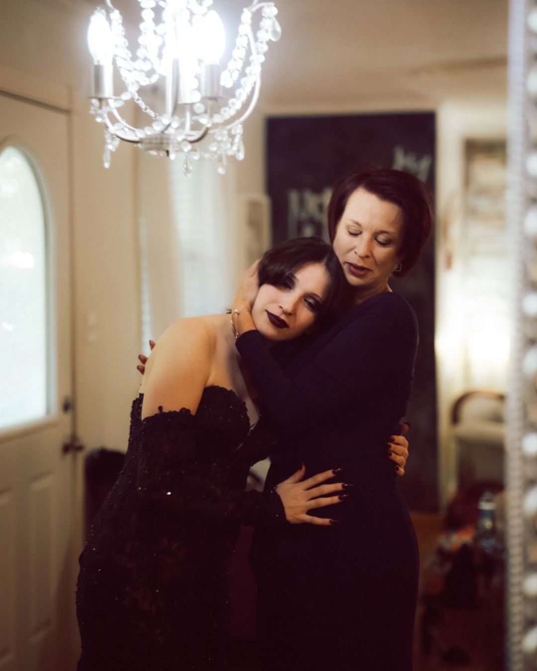 Two women in black dresses sharing a warm embrace under a crystal chandelier indoors.