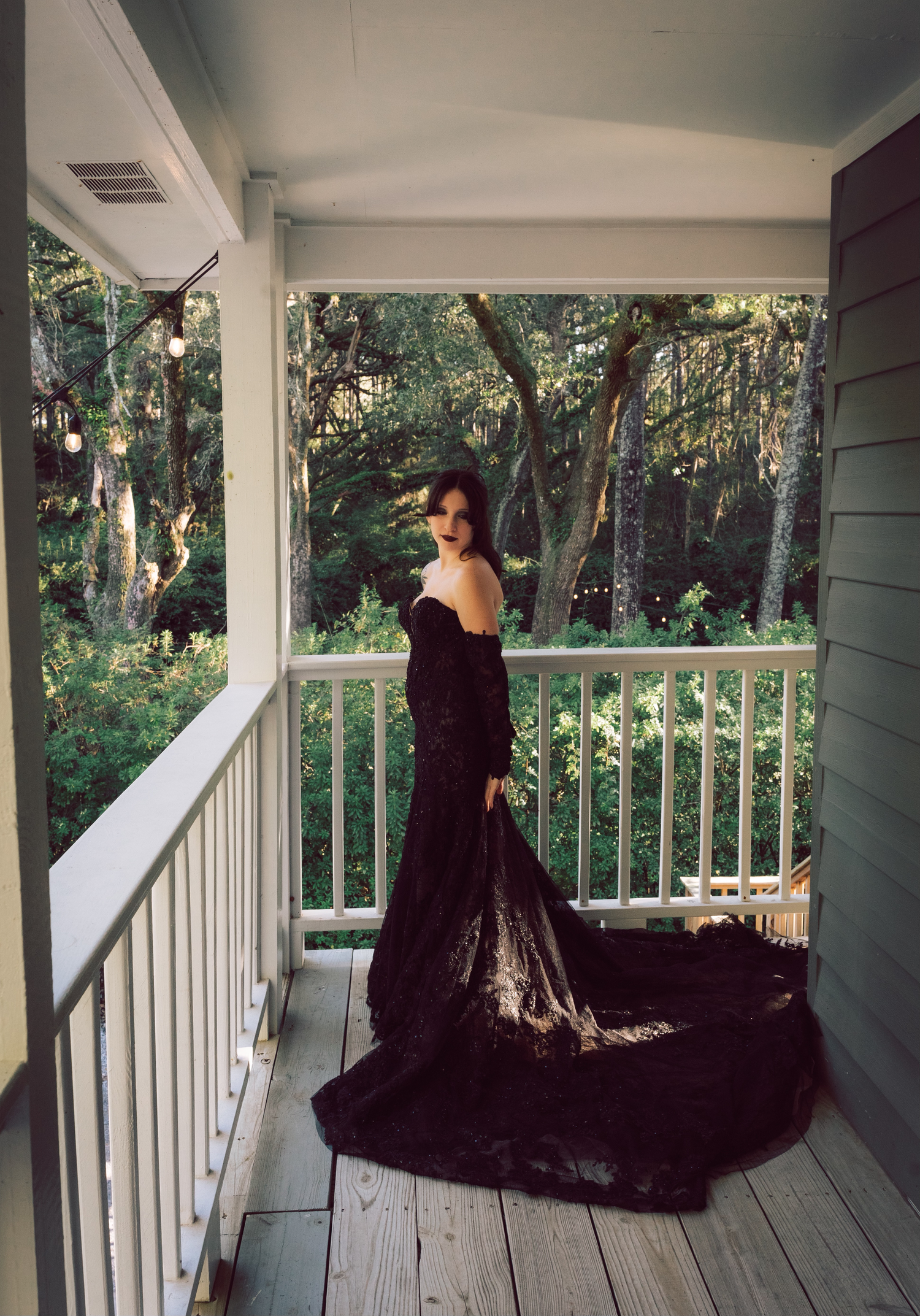Woman in elegant black off-shoulder gown with long train standing on a wooden porch surrounded by trees.