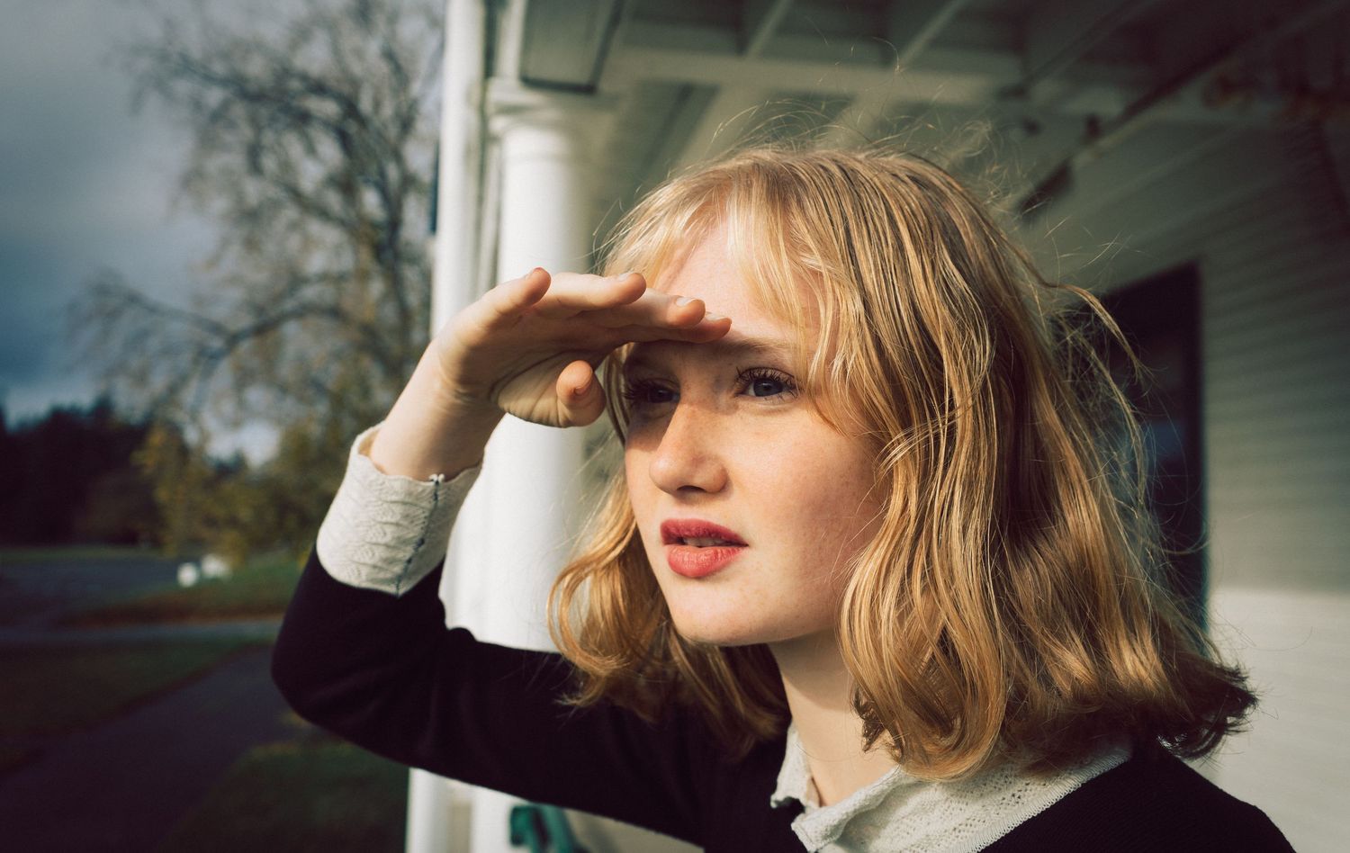 Young blonde woman shielding her eyes from the sun while standing outdoors near a white building.