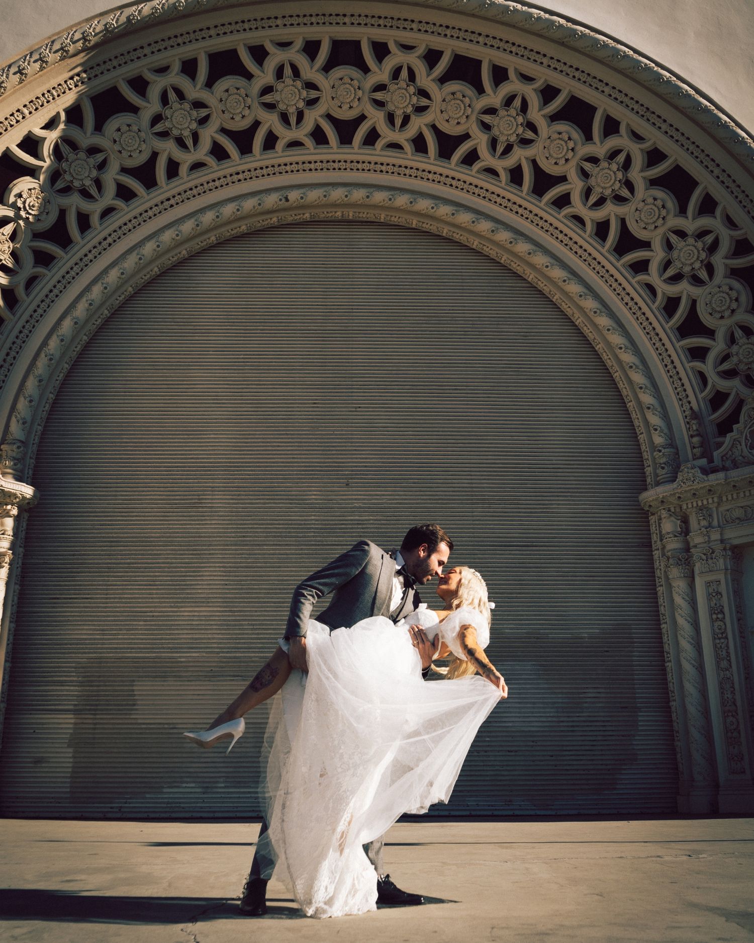 Wedding couple sharing a romantic dip kiss beneath an ornate stone arch in dramatic sunlight.