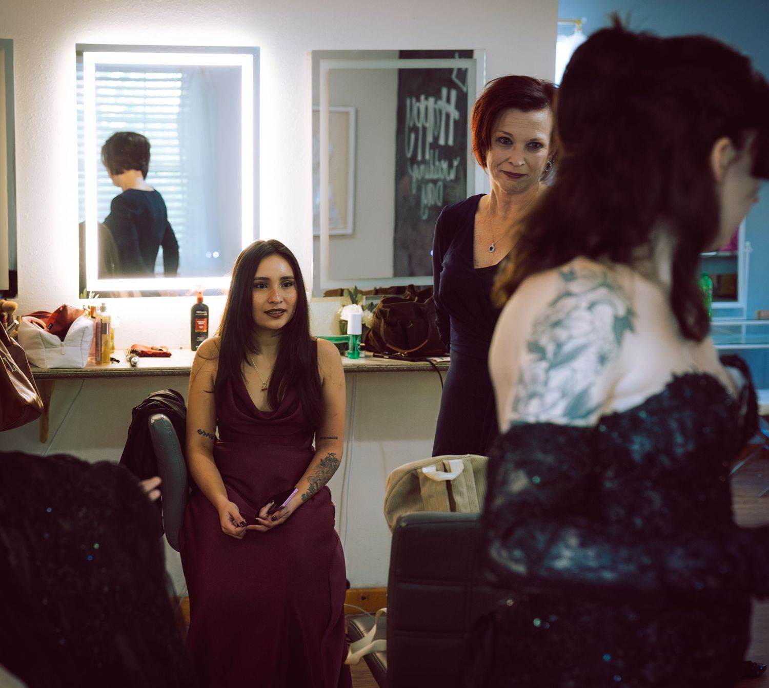 Three women in formal attire gather in a dressing room with mirrors and vanity lights.