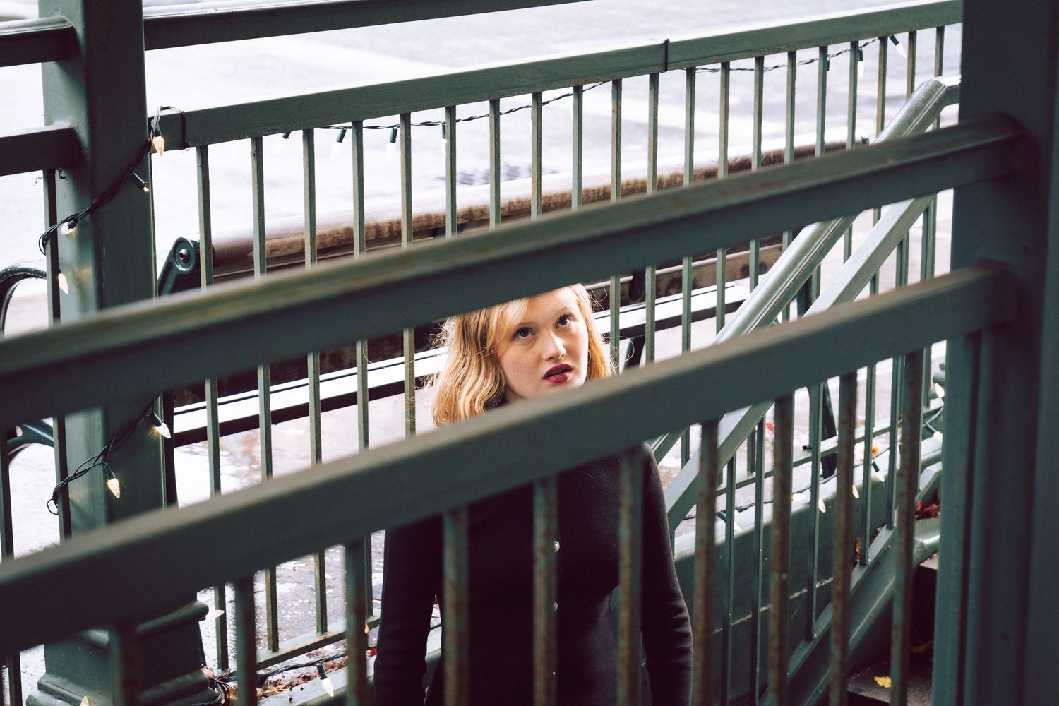 Young blonde woman peering through green metal staircase railings in an urban setting.
