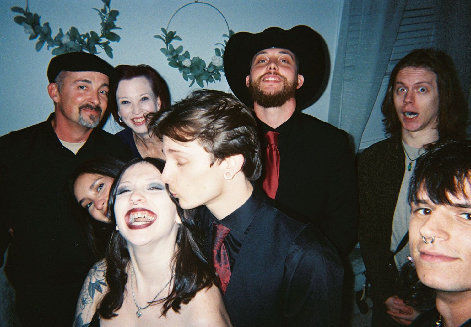 Group of wedding guests posing together indoors, groom kissing bride on cheek, cowboy hat visible.