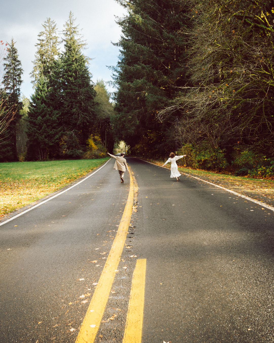 Two people dancing joyfully on a tree-lined road with yellow center lines in autumn forest setting.