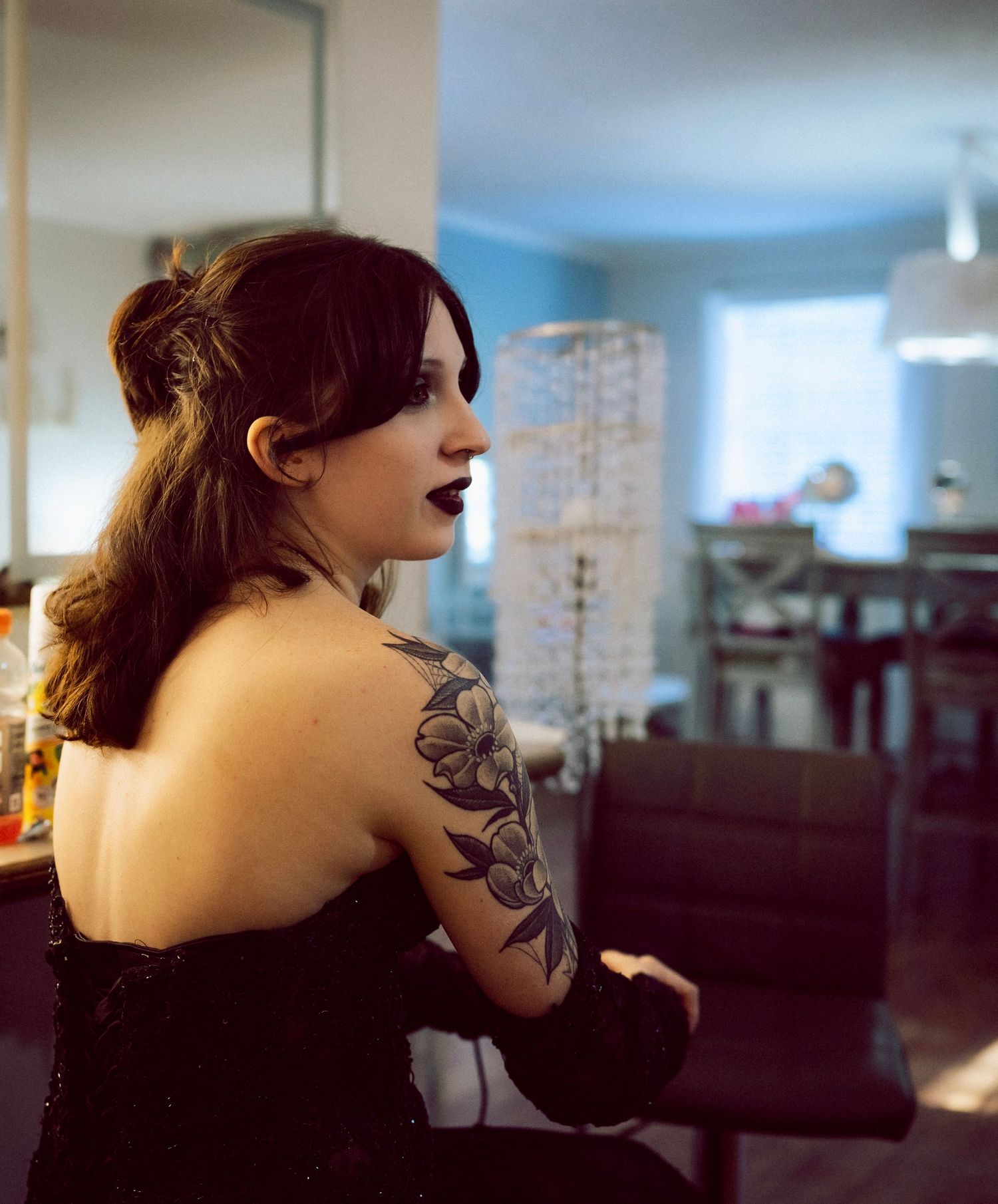 Dark-haired woman with floral shoulder tattoo wearing a black strapless dress in a home interior.