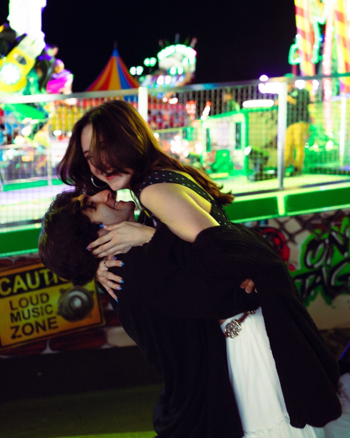 Romantic couple sharing a dip kiss at a colorful carnival fairground at night.