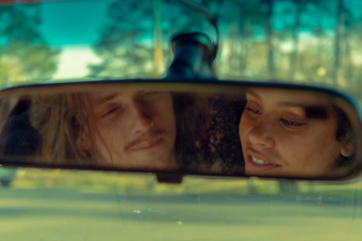 Reflection of a couple in a rearview mirror, sharing a tender moment while driving through pine trees.
