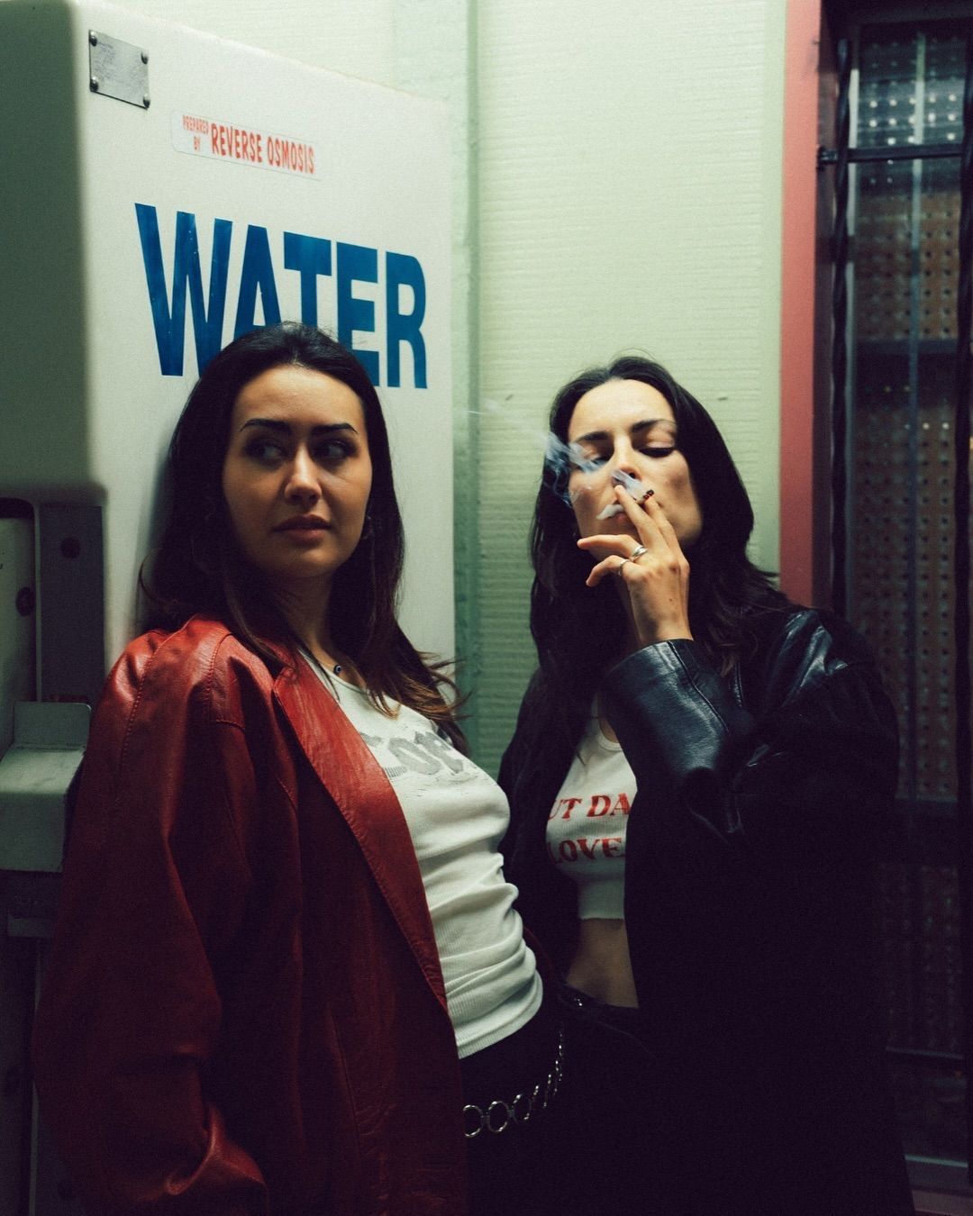 Two young women pose near a water vending machine at night, one smoking a cigarette.