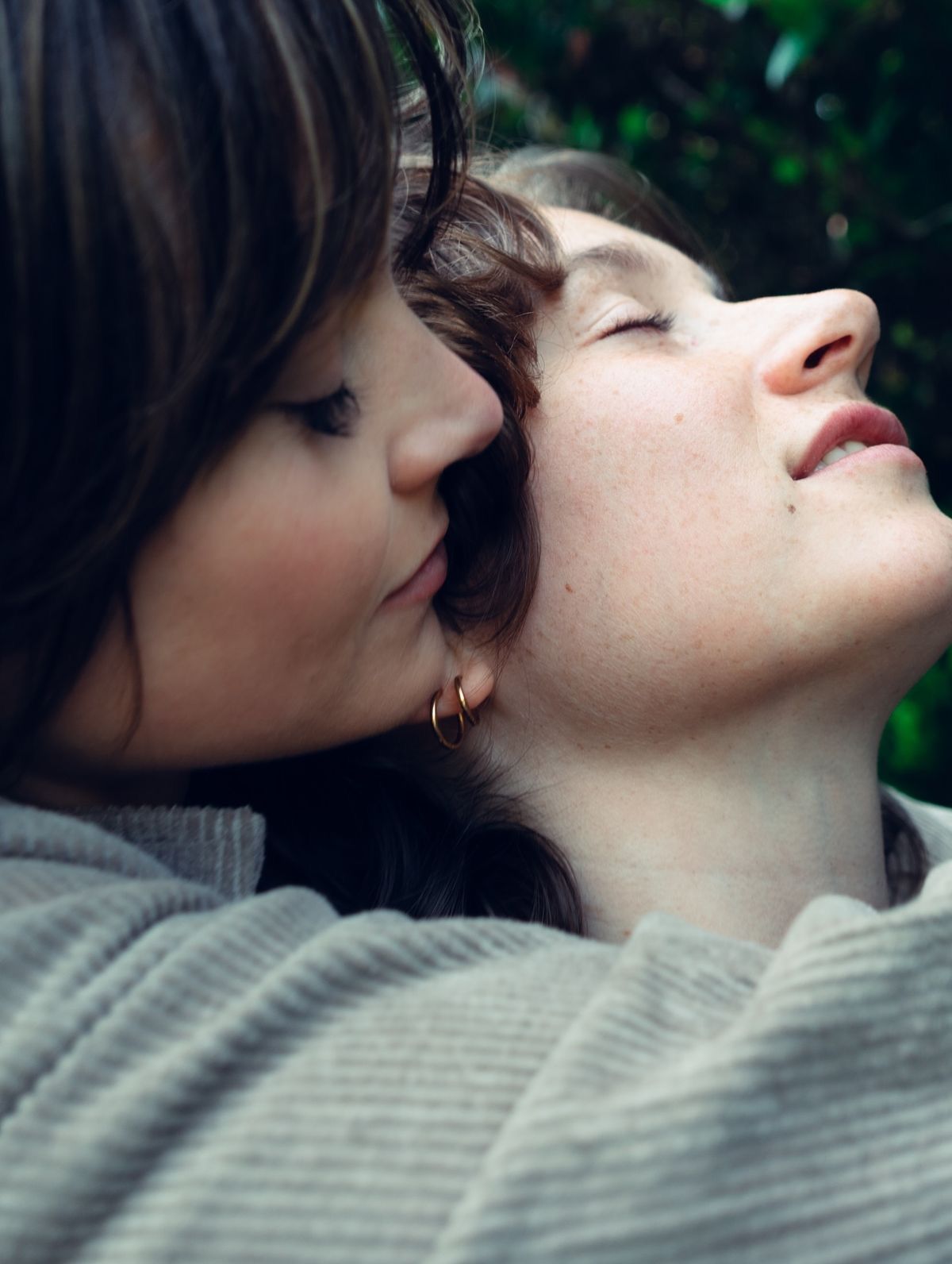 Two women share an intimate moment outdoors, one gently kissing the other's cheek near green foliage.