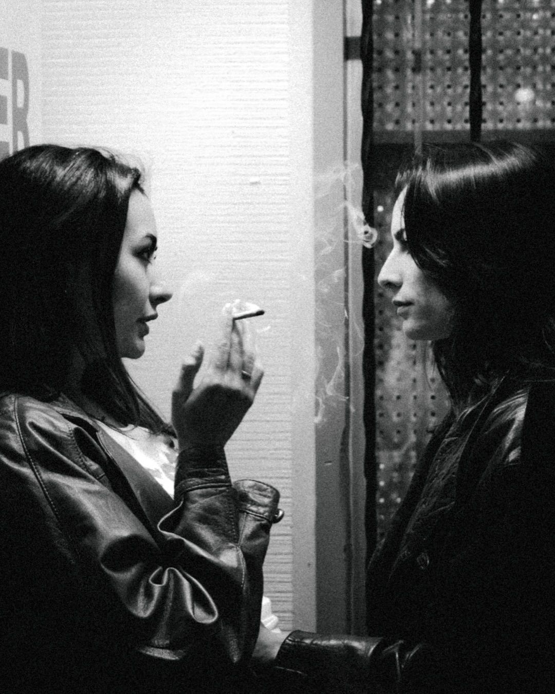 Two women in leather jackets face each other in black and white, one smoking a cigarette.