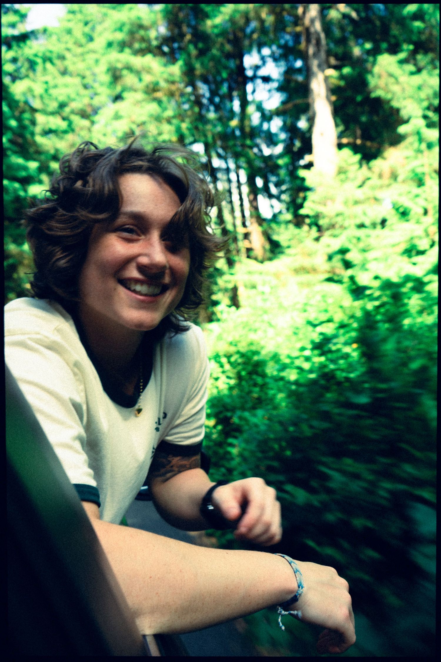 Smiling young person with curly hair leaning out car window surrounded by lush green forest.
